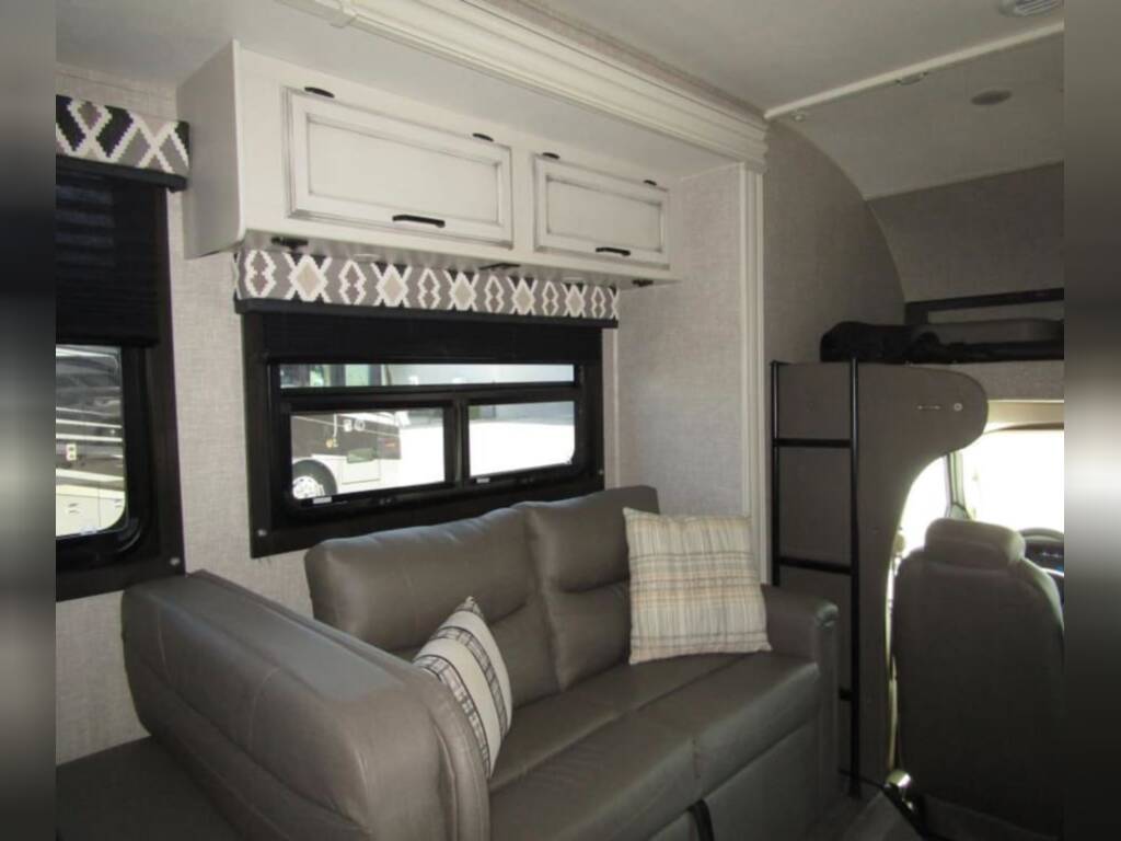 Spacious rv rental interior featuring a comfy sofa, overhead cabinets, large windows, and an over-cab bunk for sleeping.. Jayco Redhawk 2022