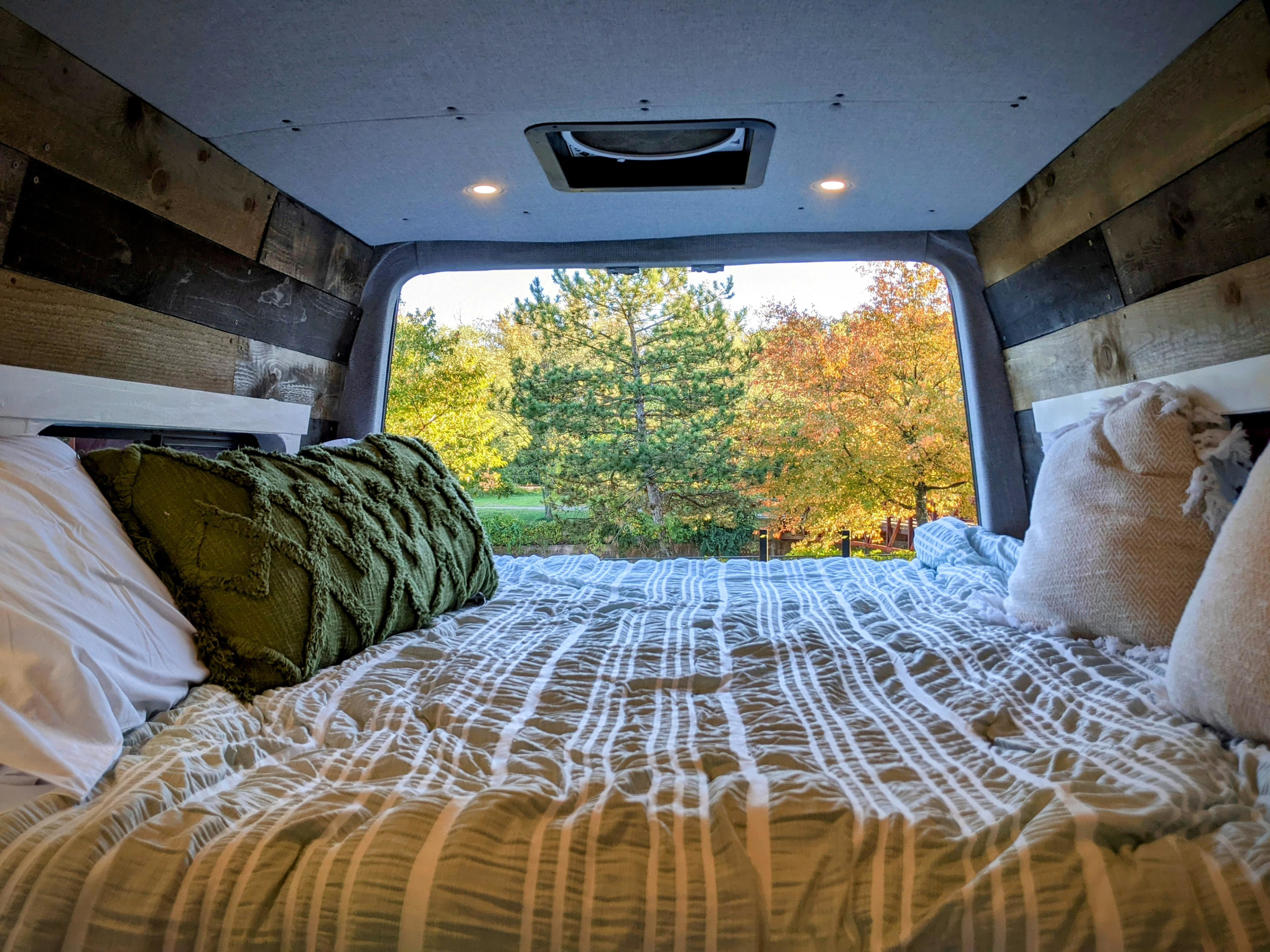 Cozy RV rental camper van interior. A bed with a green pillow and striped comforter offers a serene view of autumn trees.. Mercedes-Benz Sprinter 2008