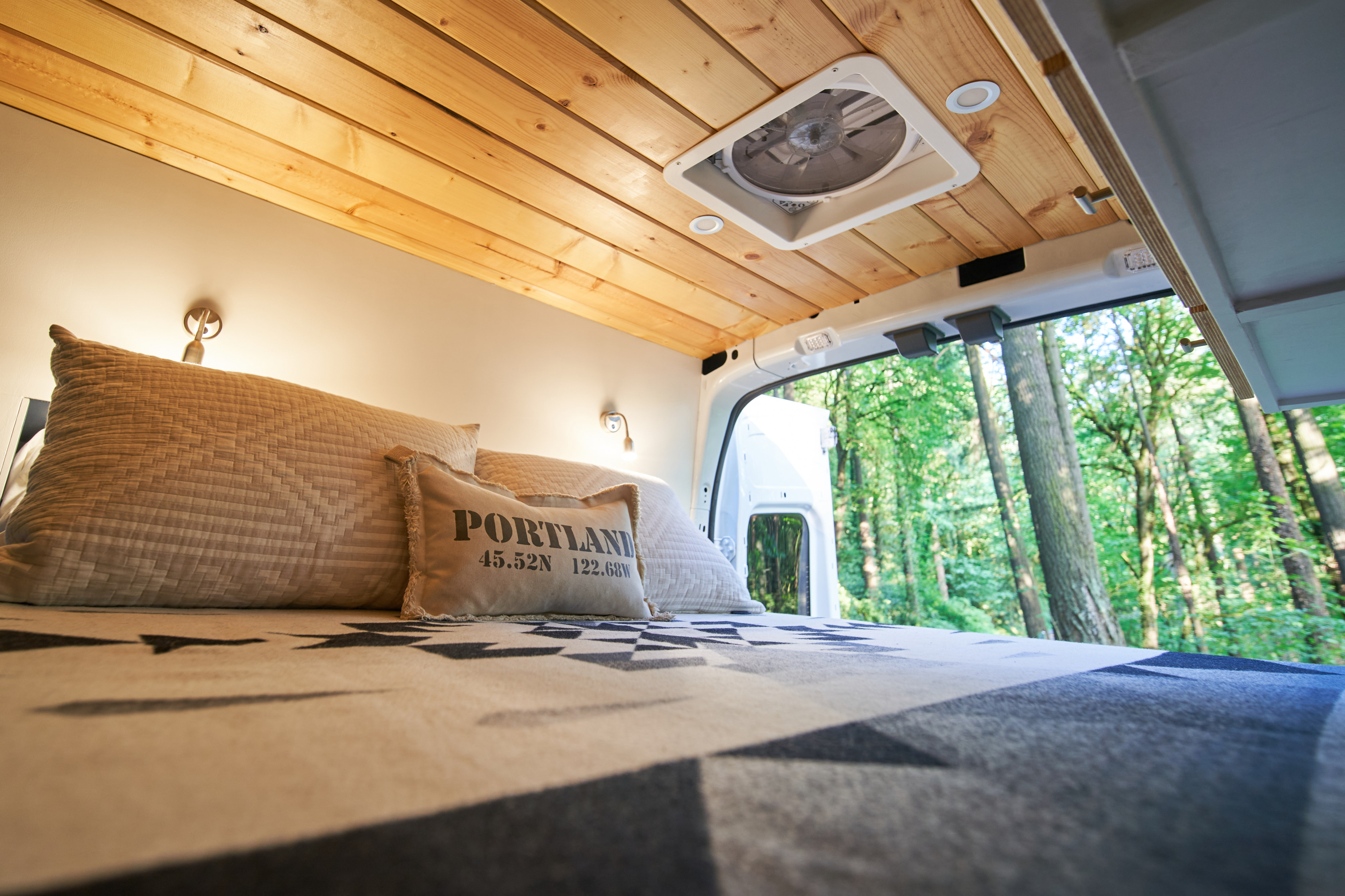 Relaxing RV rental bed with a 'Portland' pillow, wooden ceiling, and serene forest view from the open rear doors.. Ford Transit 2021
