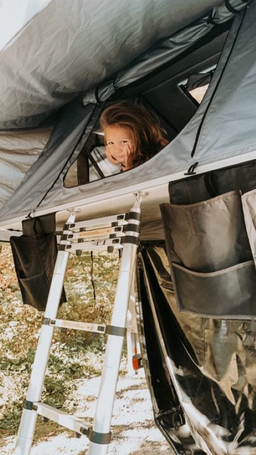 Child smiles from a rooftop tent on an RV rental, ready for adventure. Ladder and storage are visible.. Rooftop Camp S'PA Lourd 2024
