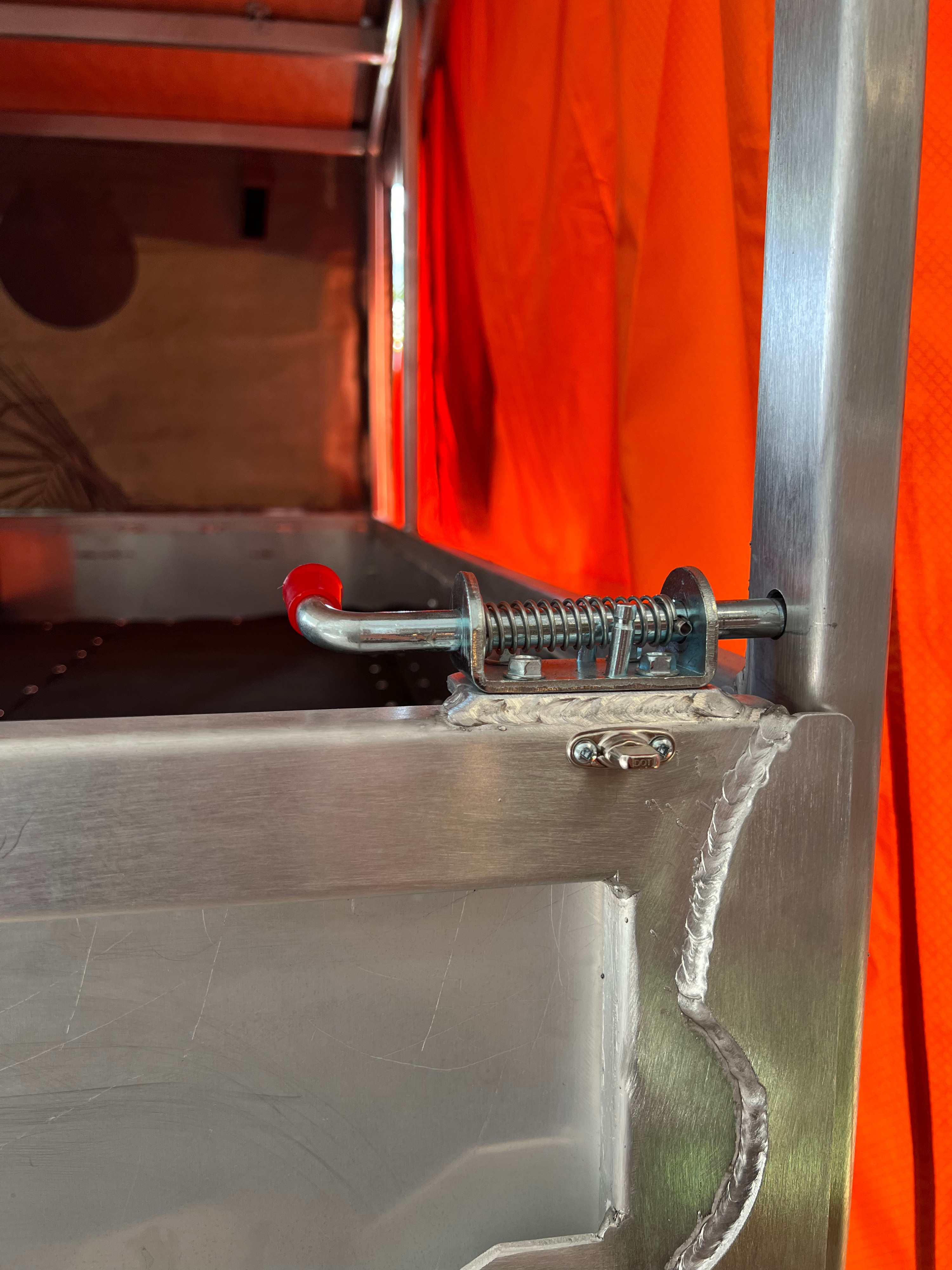 RV rental security: Detailed view of a heavy-duty metal pin latch on a camper's door frame, with orange interior backdrop.. Rooftop Camp S'PA lourd 2024