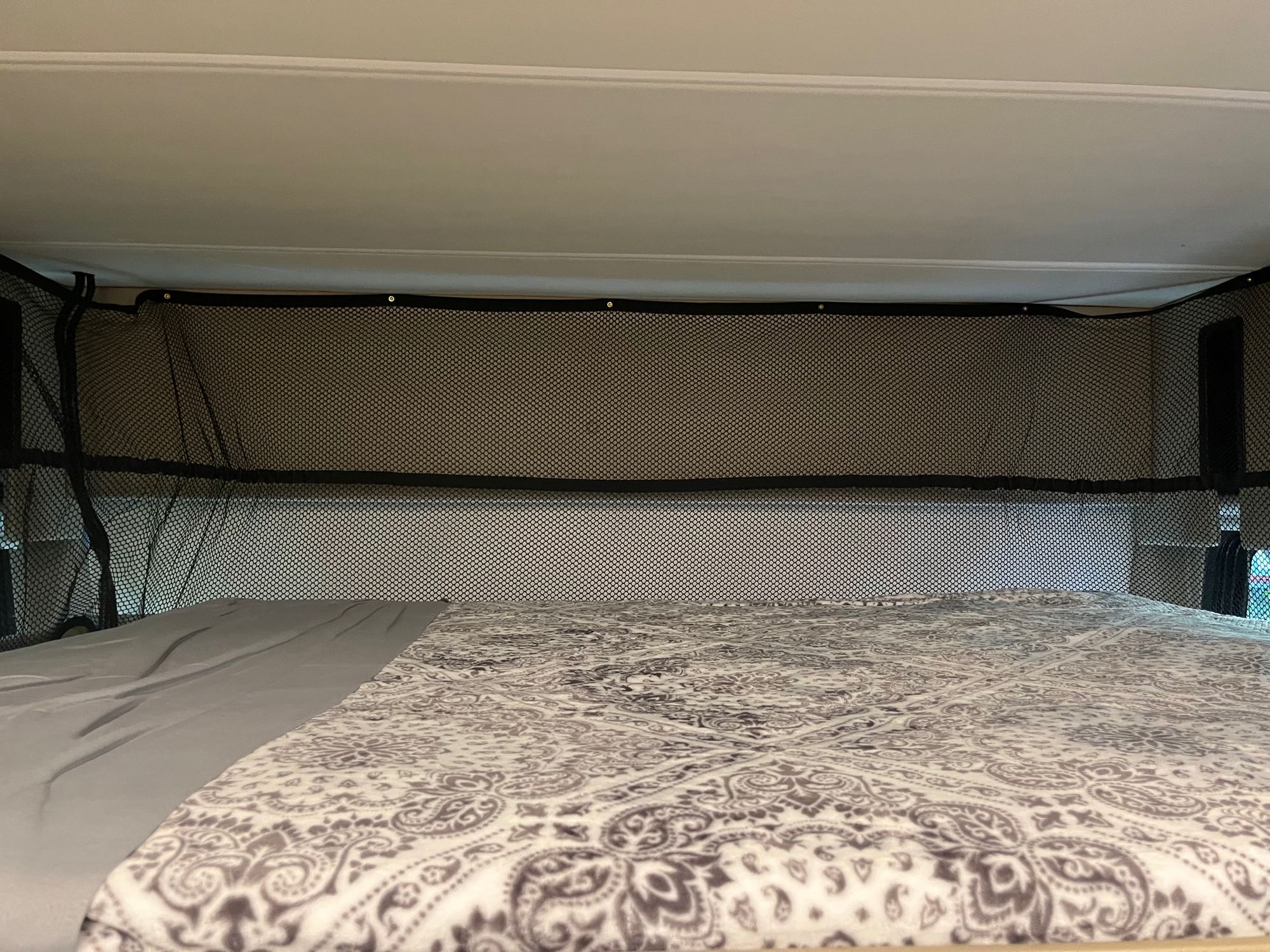 Inside an RV rental: a cozy bed with a patterned blanket and overhead mesh safety net in a sleeping bunk.. Thor Challenger 2021