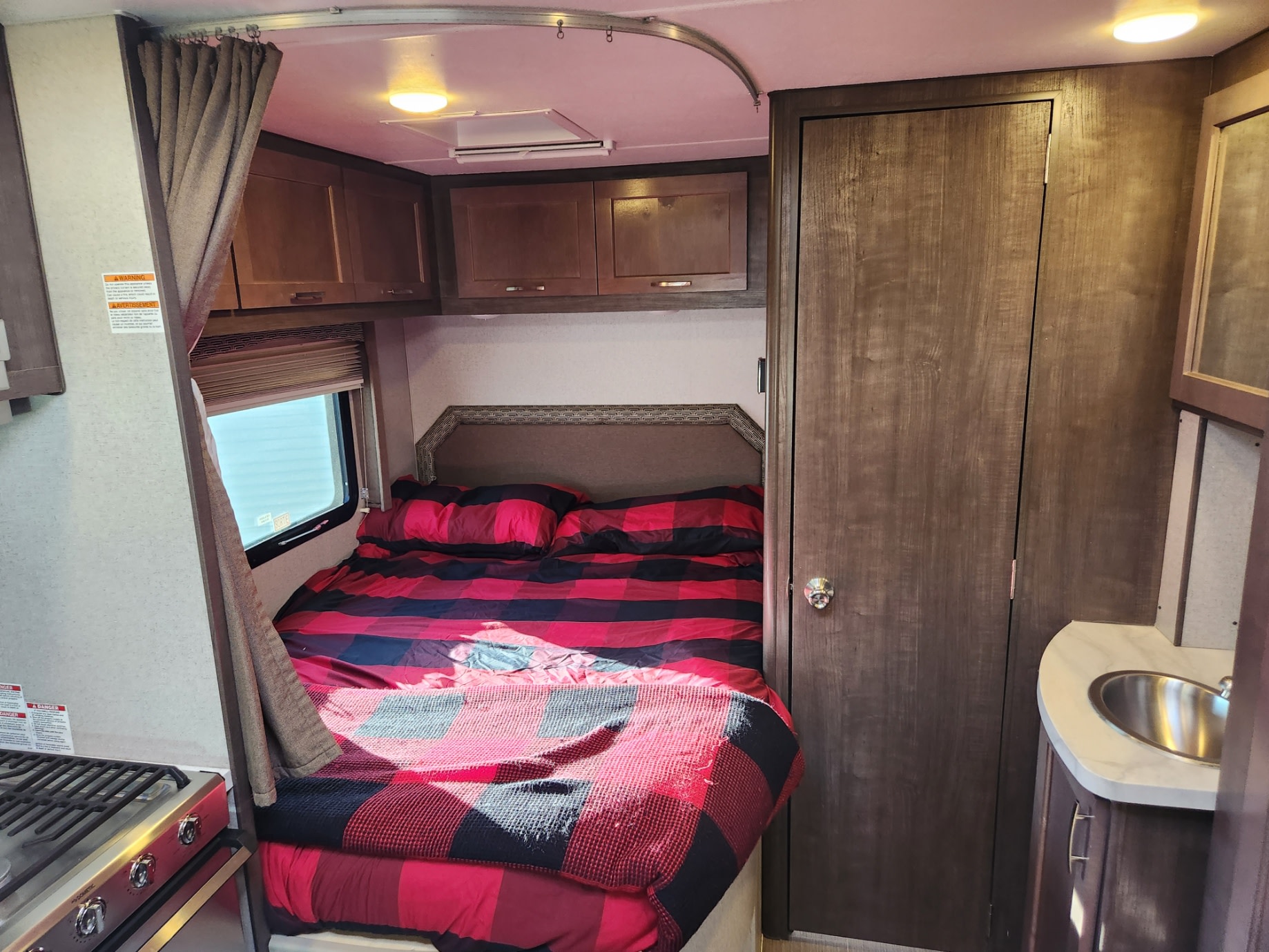 Inside an RV rental, showing a cozy bedroom with red plaid bedding, small kitchen with stove, and a sink.. Adventurer 24DS 2020