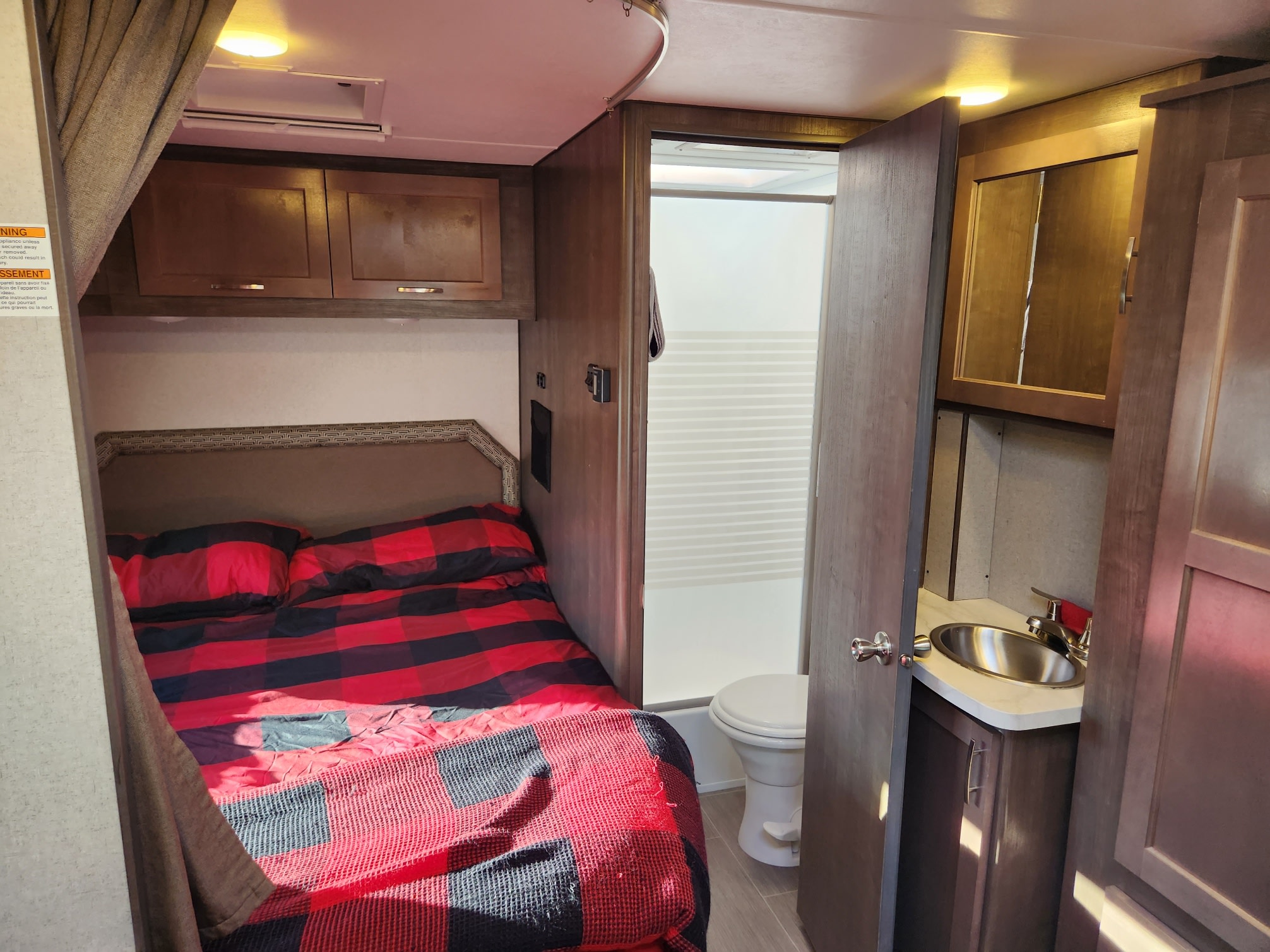 Cozy RV rental interior showing a bedroom with a red plaid bed and an adjacent compact bathroom with toilet and sink.. Adventurer 24DS 2020