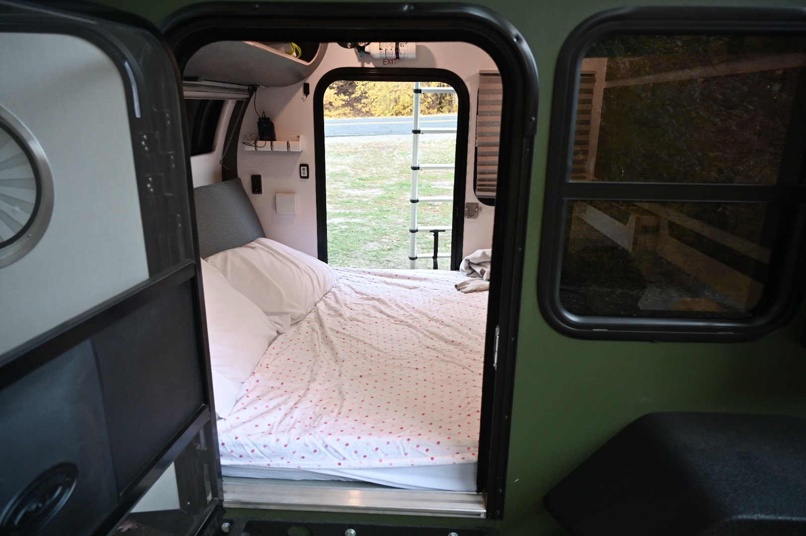 Inside an RV rental: a compact sleeping area with a polka dot comforter. The open rear hatch reveals a grassy outdoor view.. bean bean trailer 2021
