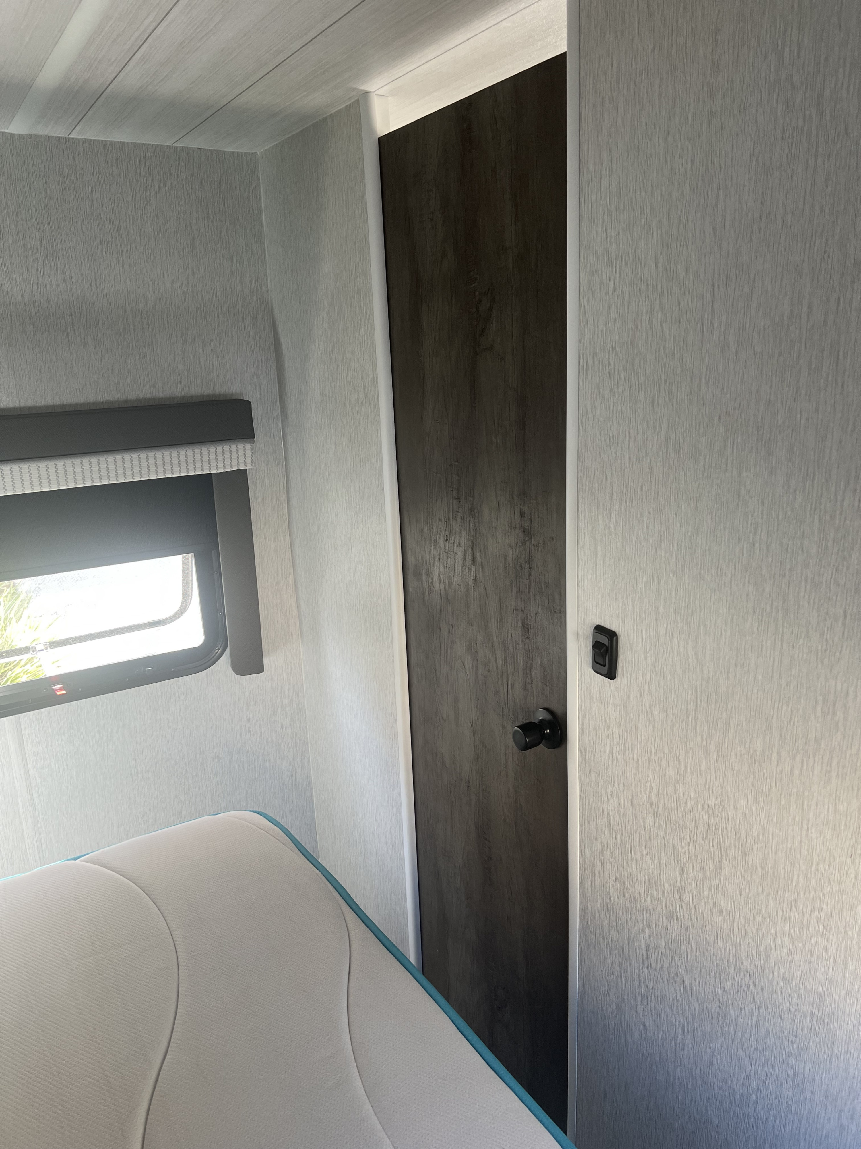 Inside an RV rental: a compact bedroom with a dark door, sleek gray textured walls, and a window by the bed.. Forest River Salem Cruise Lite 2022