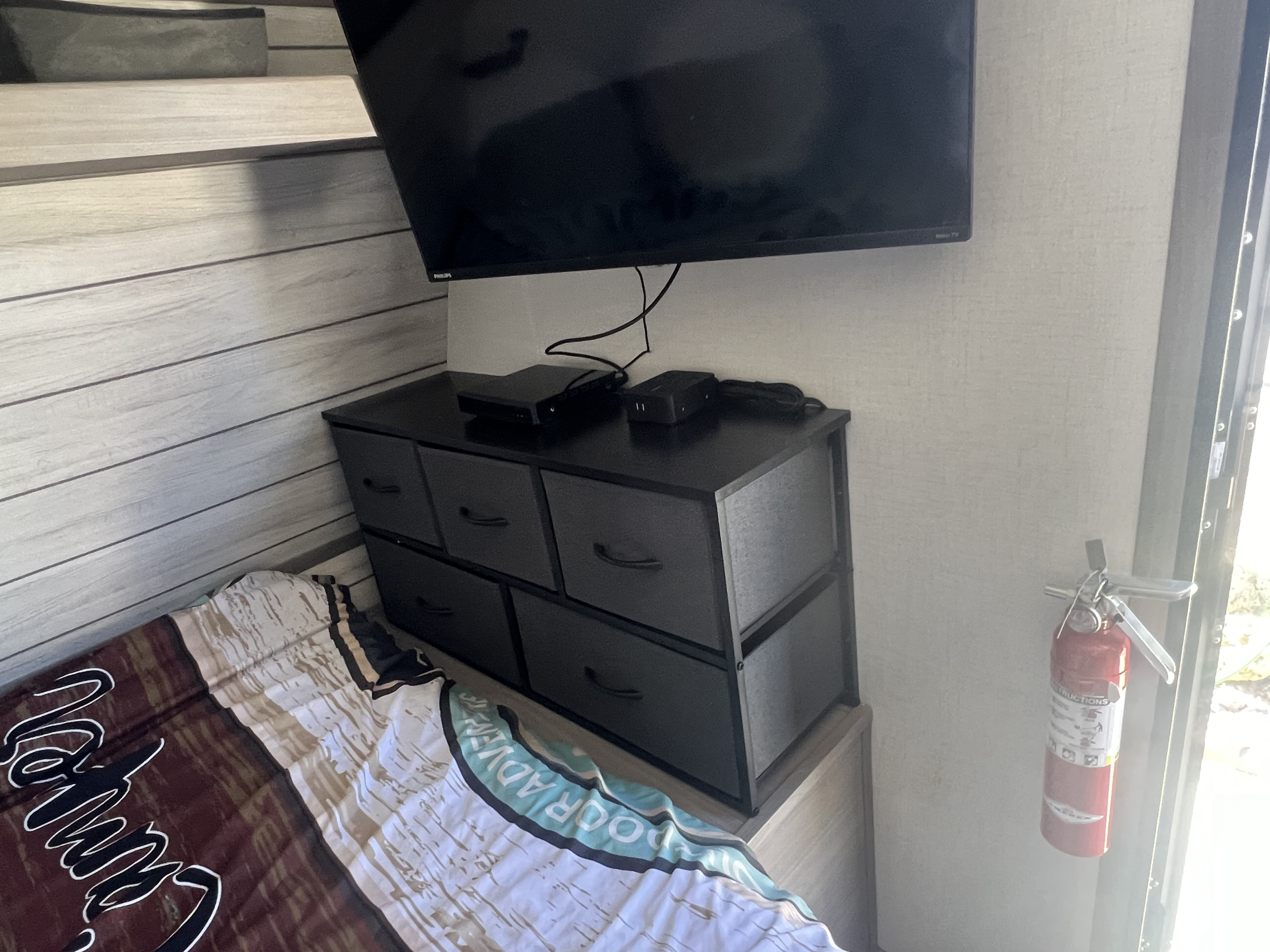 Cozy RV rental bedroom featuring a wall-mounted TV, fabric dresser, and safety fire extinguisher next to the bed.. Keystone RV Coleman 2024