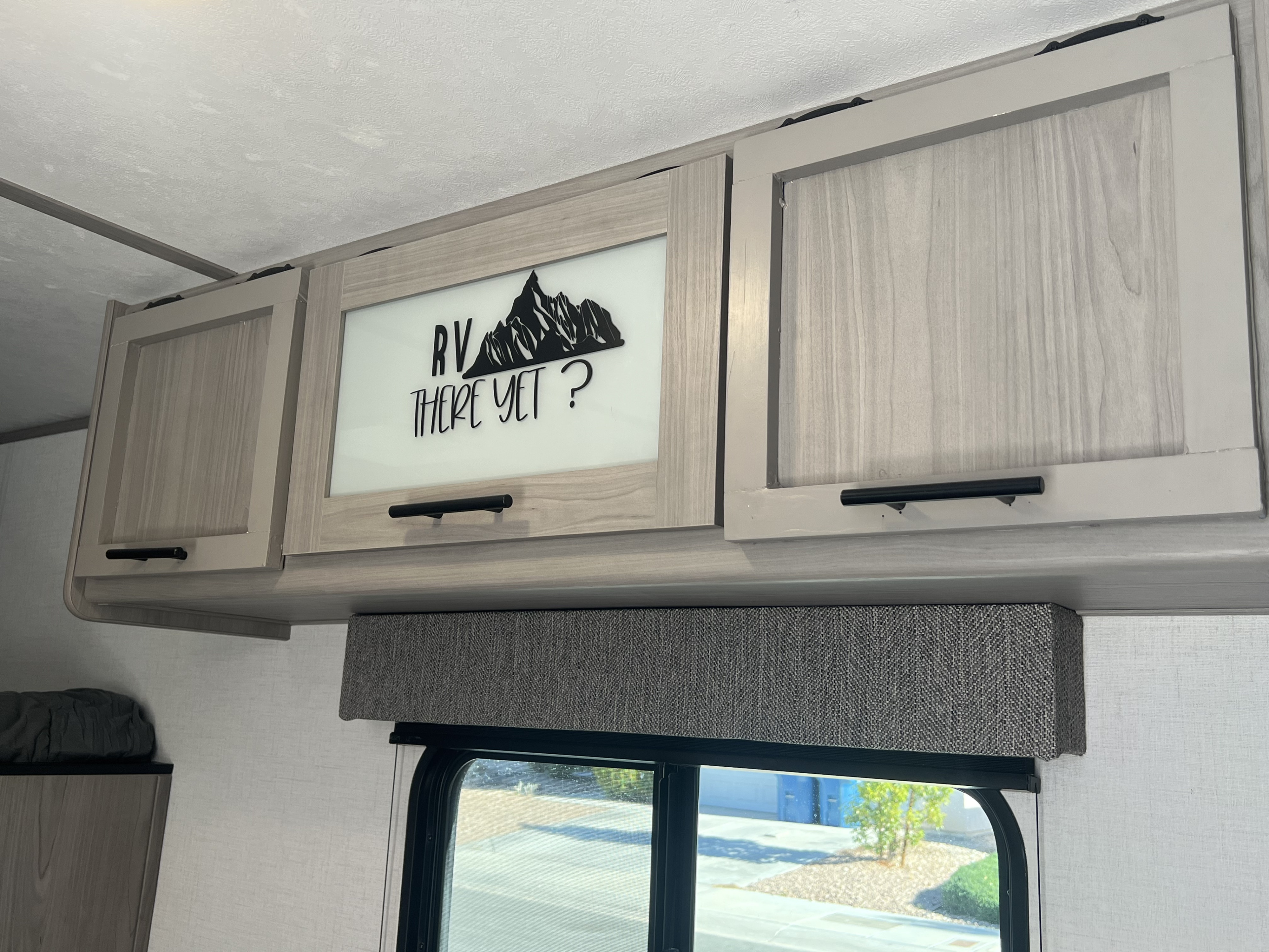 RV rental interior: Light wood overhead cabinets, including one with "RV THERE YET?" and a mountain graphic, above a window with a fabric valance.. Keystone RV Coleman 2024
