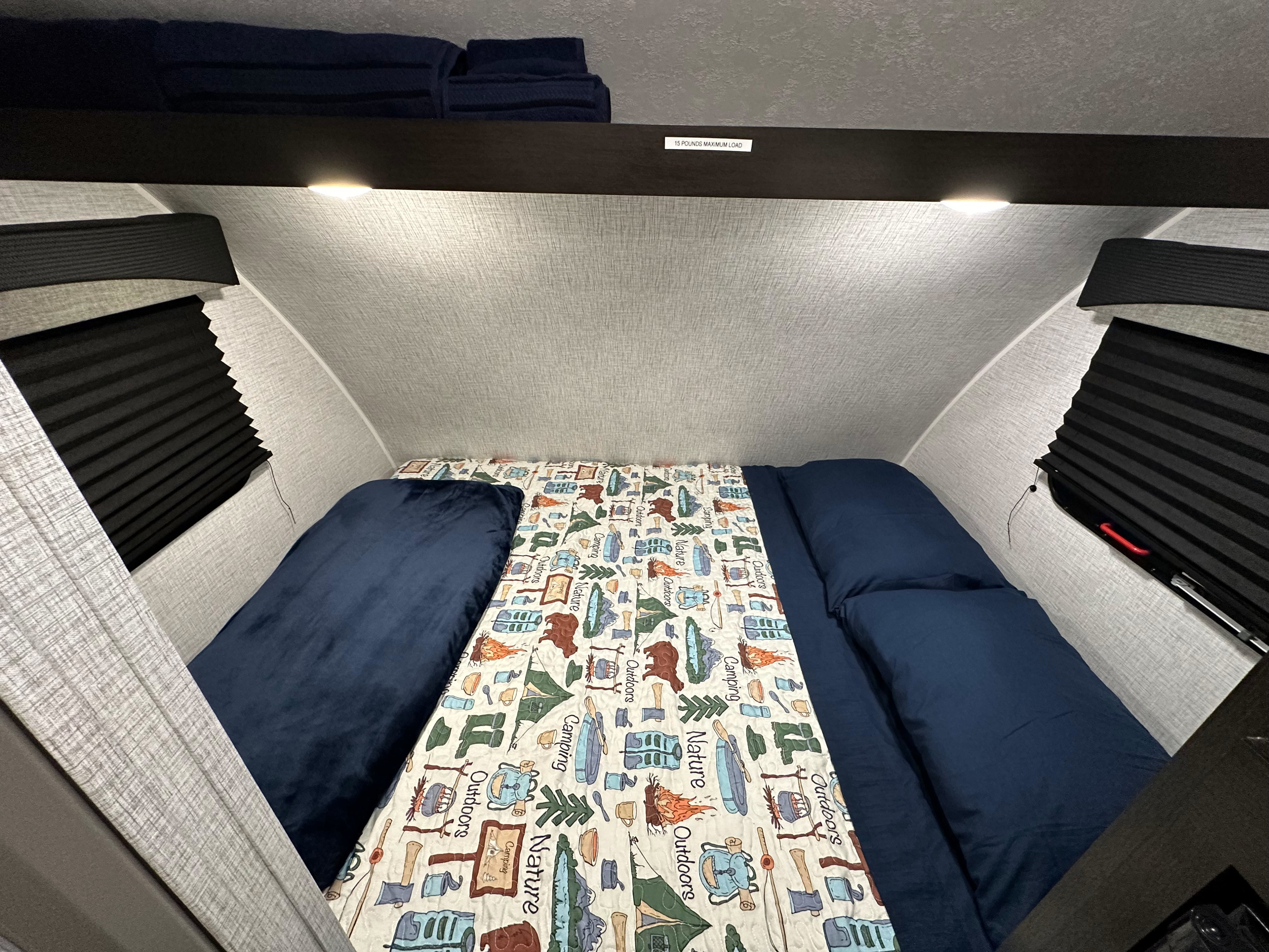 RV rental's cozy sleeping area: a bed with a camping-themed quilt, navy bedding, and overhead storage with a 15 lb limit.. Braxton Creek Bushwhacker Plus 2023