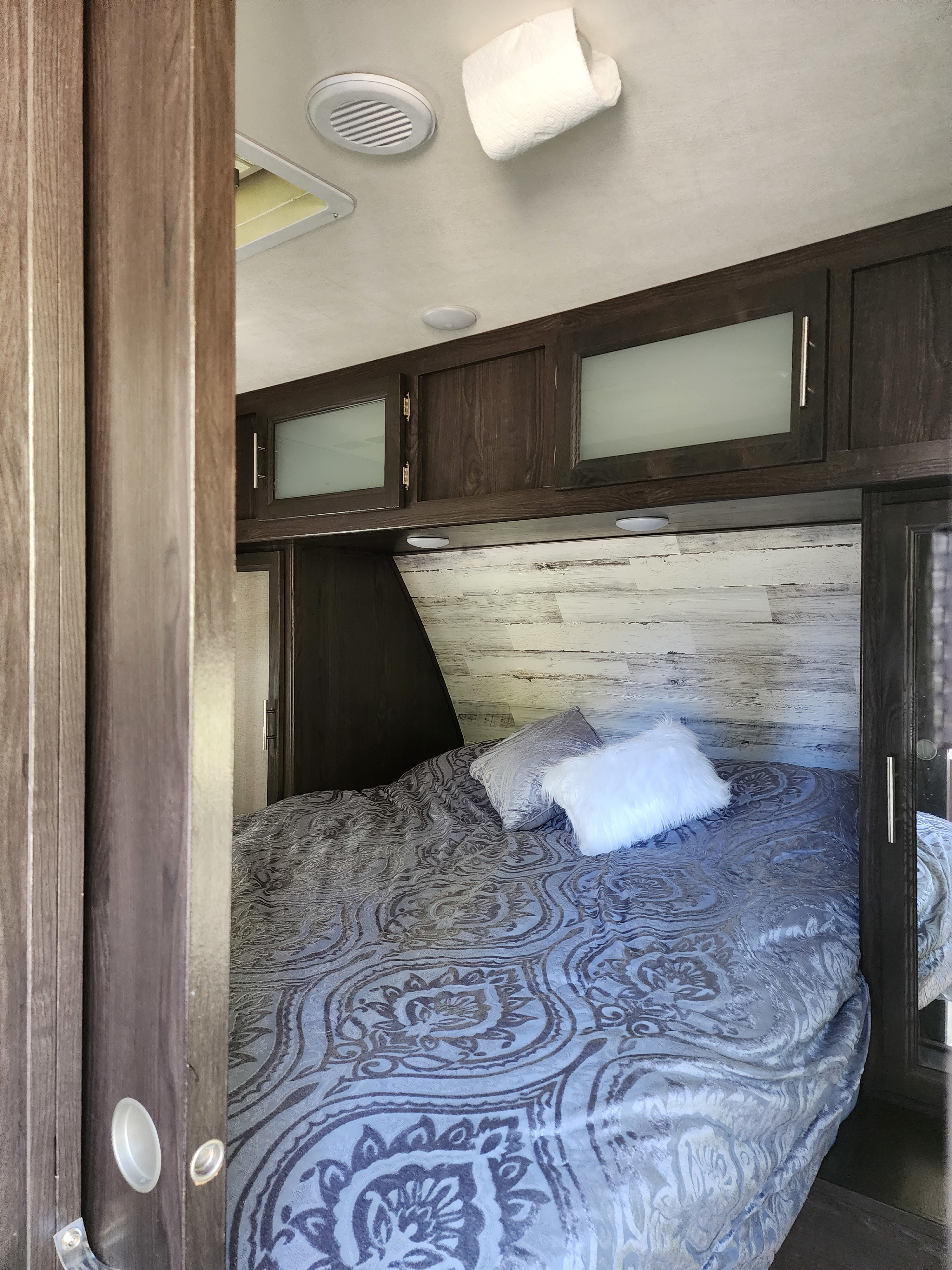 RV rental bedroom interior: Features a bed with patterned linens, a distressed wood headboard, and dark wood overhead cabinets.. Dutchmen Kodiak Ultra Lite 2019