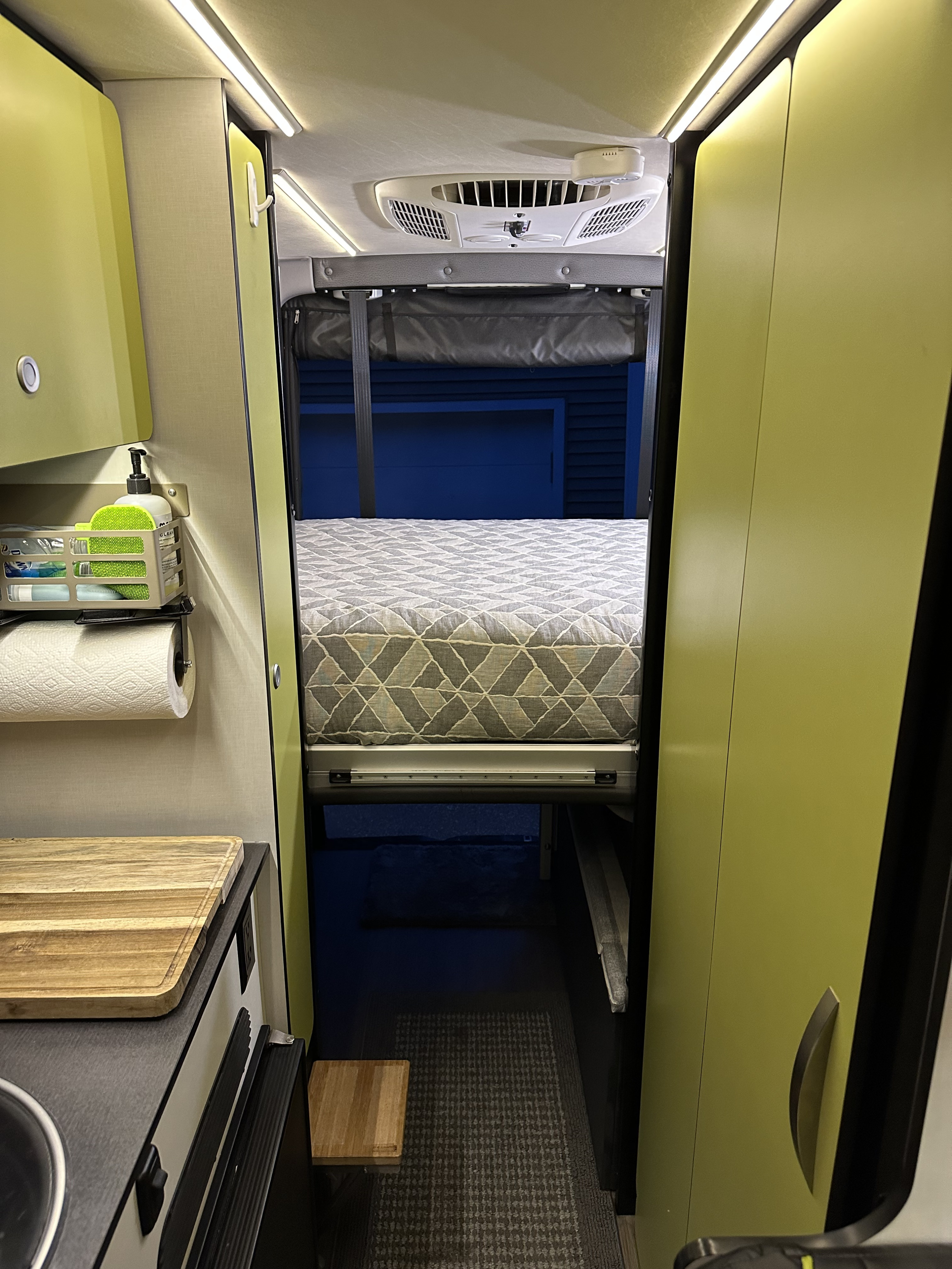 A bright rv rental interior, showcasing a convertible bed, compact kitchen with a sink, and lime-green cabinets, all within a narrow aisle.. Winnebago Revel 2020