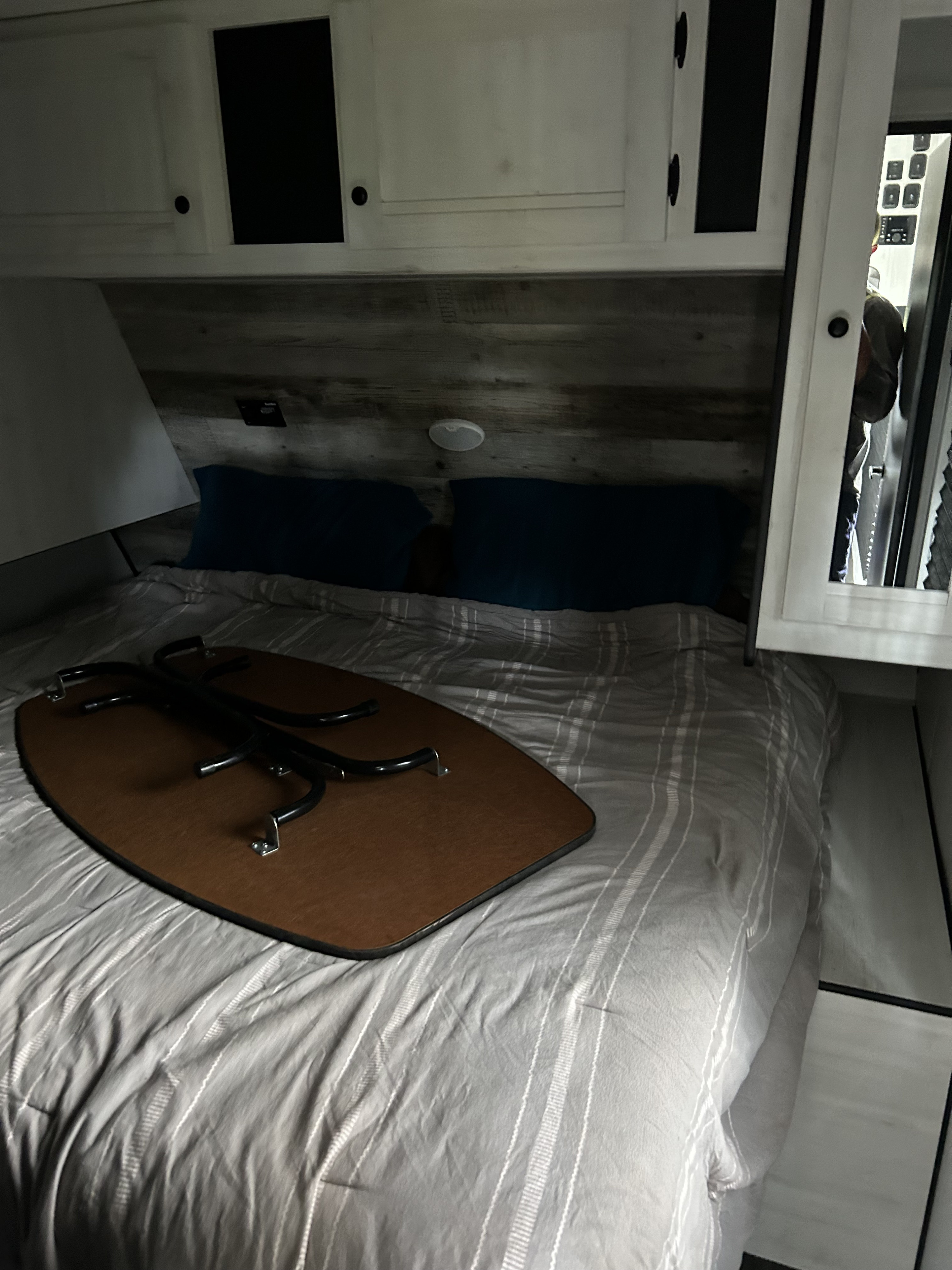 RV rental bedroom with a bed, striped bedding, blue pillows, and a portable table. Overhead cabinets are visible.. Heartland Lithium 2021