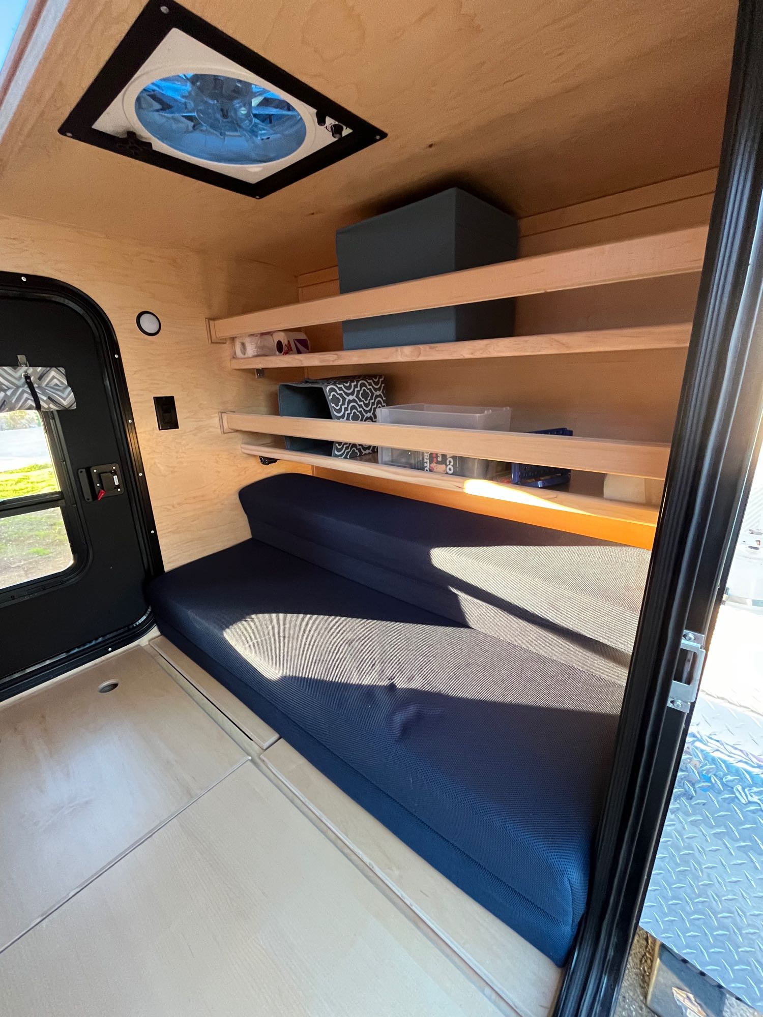 Inside a compact wood-paneled RV rental featuring a blue cushioned bed, shelves, a ceiling fan, and an entry door.. Colorado Teardrops Mount Massive 2023