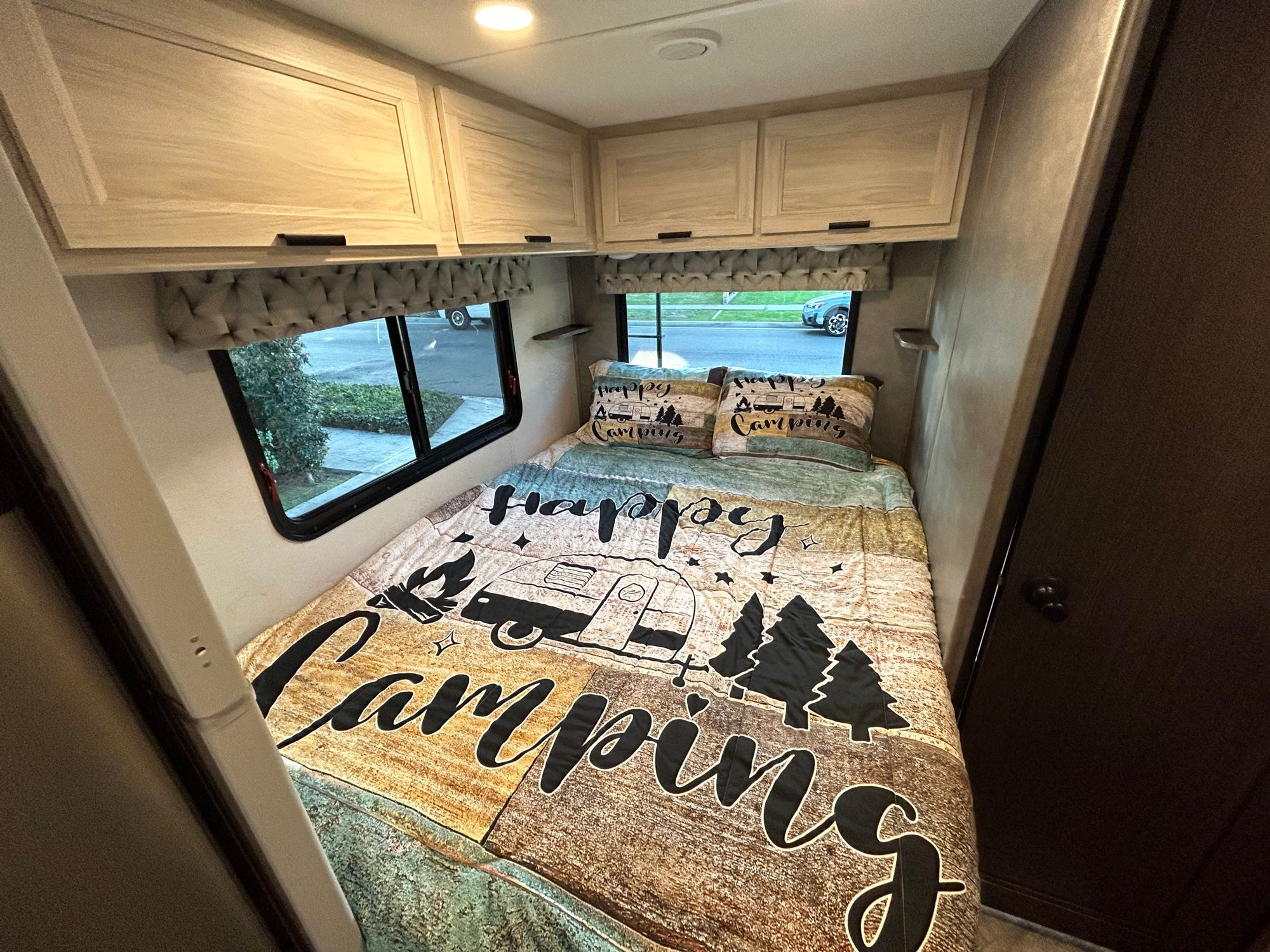 Cozy rv rental bedroom featuring a "Happy Camping" bed, overhead cabinets, and large windows. Perfect for your next adventure!. Forest River Sunseeker LE 2022