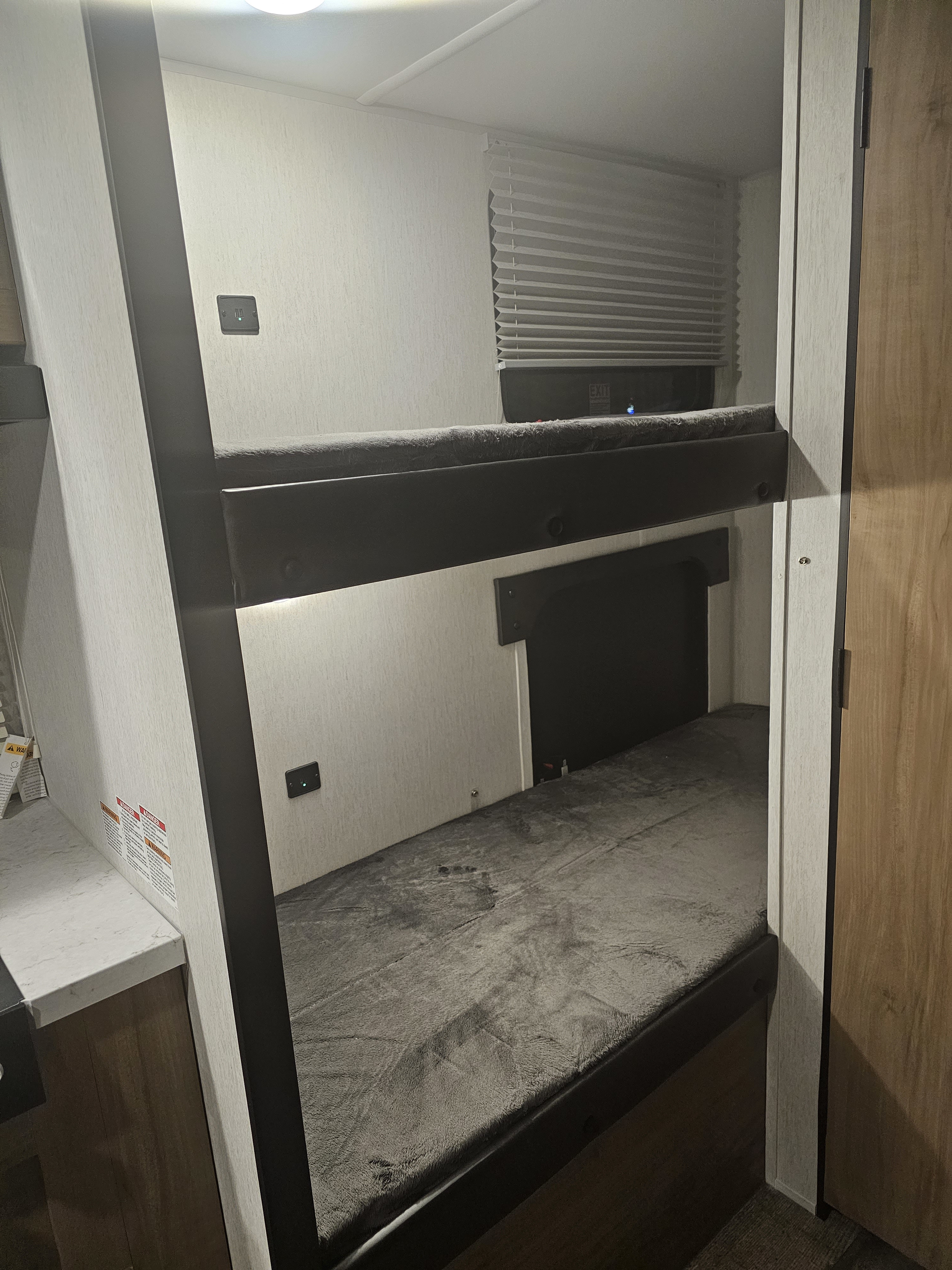 Compact RV rental interior featuring two bunk beds with soft grey blankets, wall outlets, and a window with blinds.. Forest River Cherokee Wolf Pup 2023