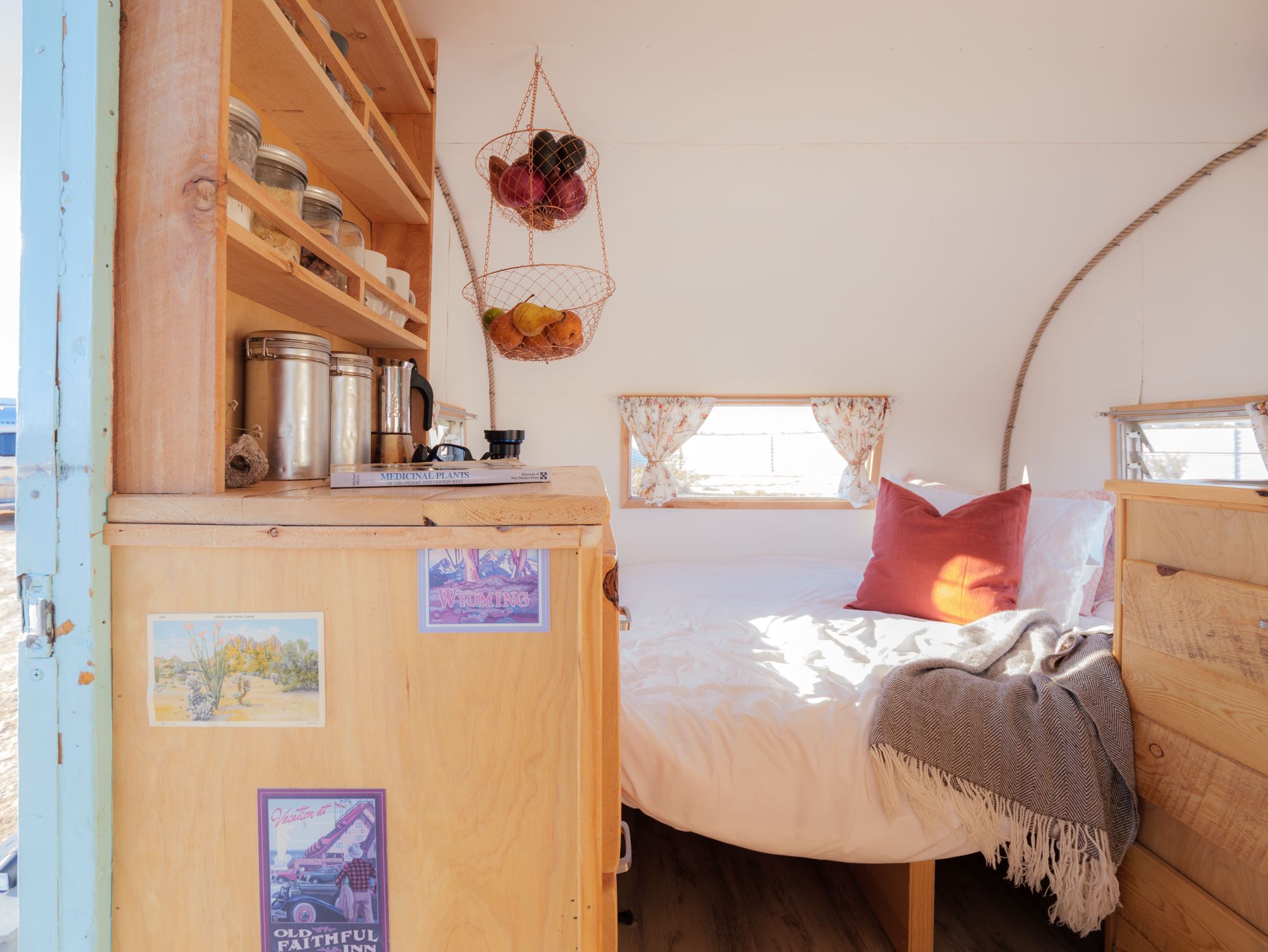 Experience comfort in this cozy rv rental: a bright bedroom with white bedding, wooden shelves, and hanging fruit baskets.. Shasta Airflyte 1962