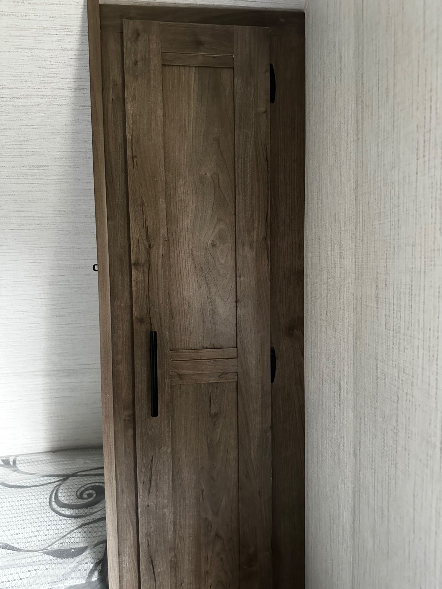 RV rental bedroom storage: Modern wood cabinet with black handles, next to a textured wall and bed.. Heartland RVs Pioneer 2024