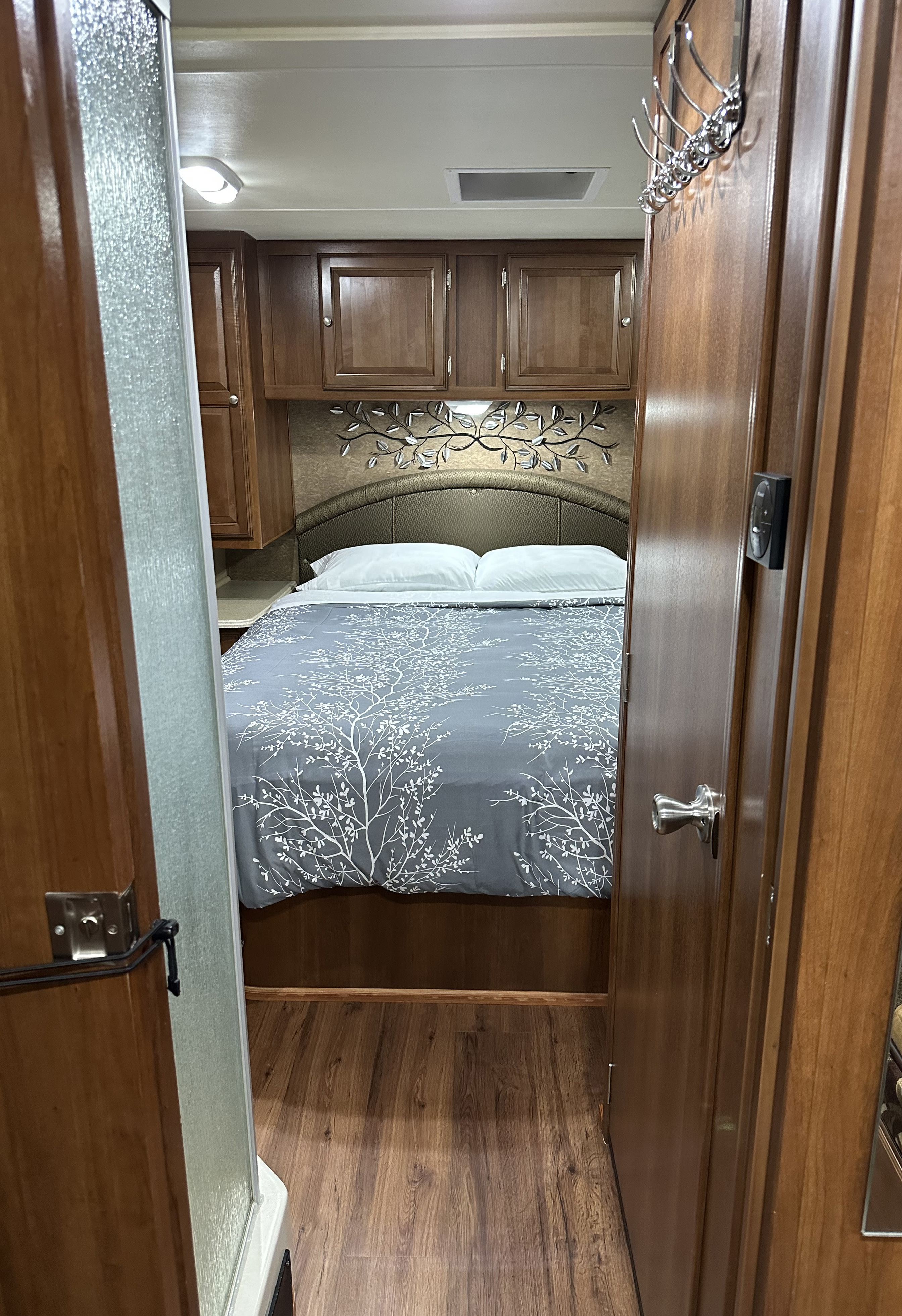 Inside an RV rental: a compact bedroom features a bed with tree-patterned duvet, overhead storage, and a nearby shower.. Forest River Coachmen Leprechaun 2015