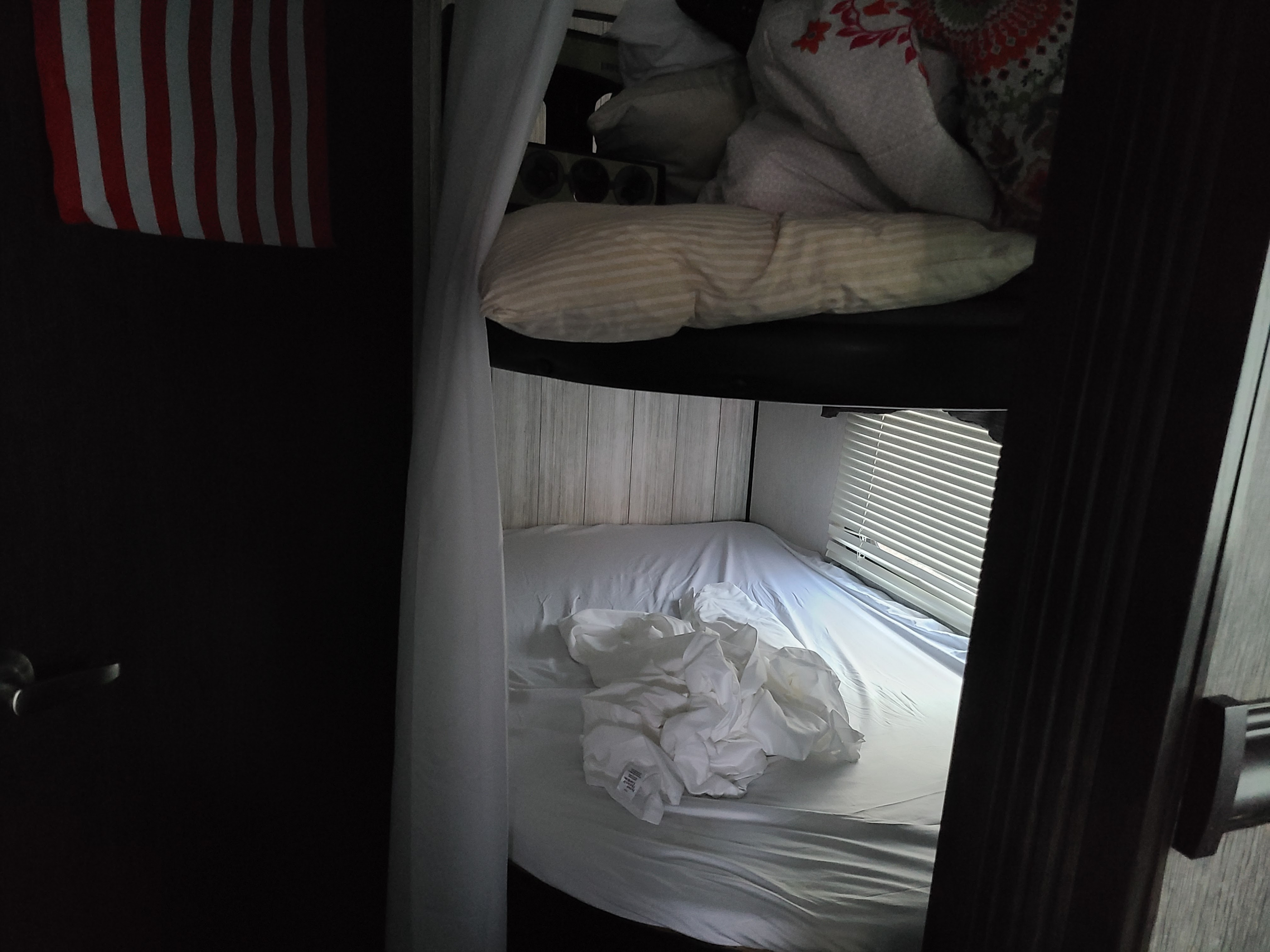 Interior view of an RV rental's bunk beds: top with patterned bedding, bottom with white sheets, blinds & flag visible.. Heartland RVs Pioneer 2019