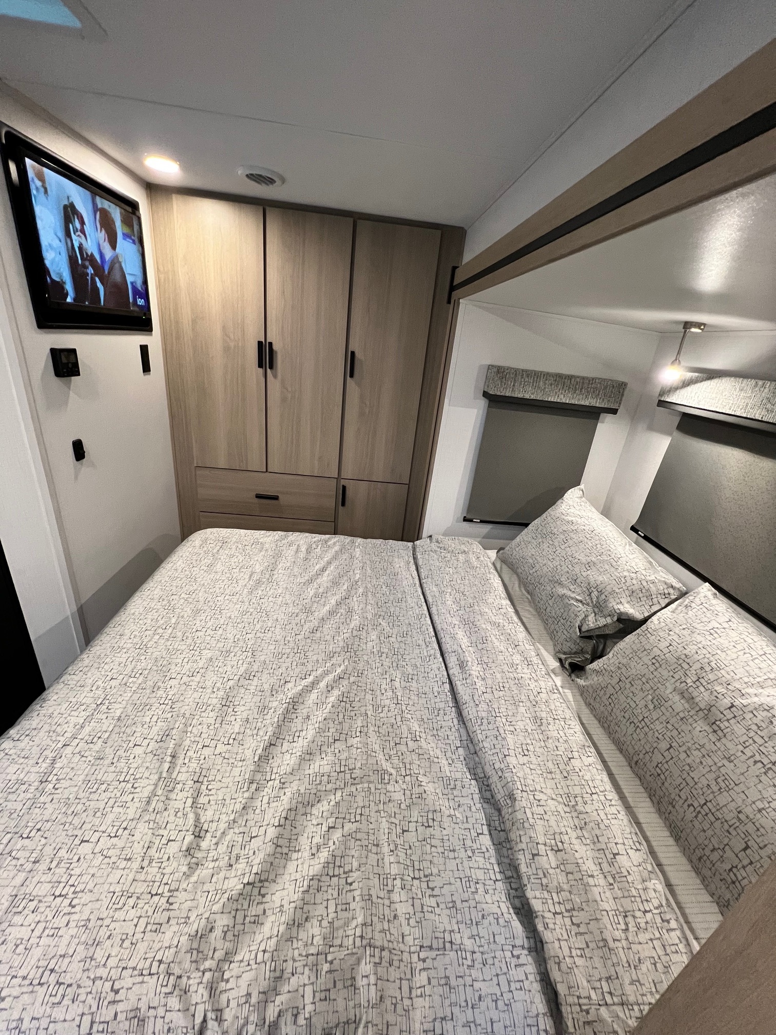 Inside an RV rental: a compact bedroom with a patterned bed, wall-mounted TV, cabinets, and shaded windows.. Forest River Sunseeker 2024