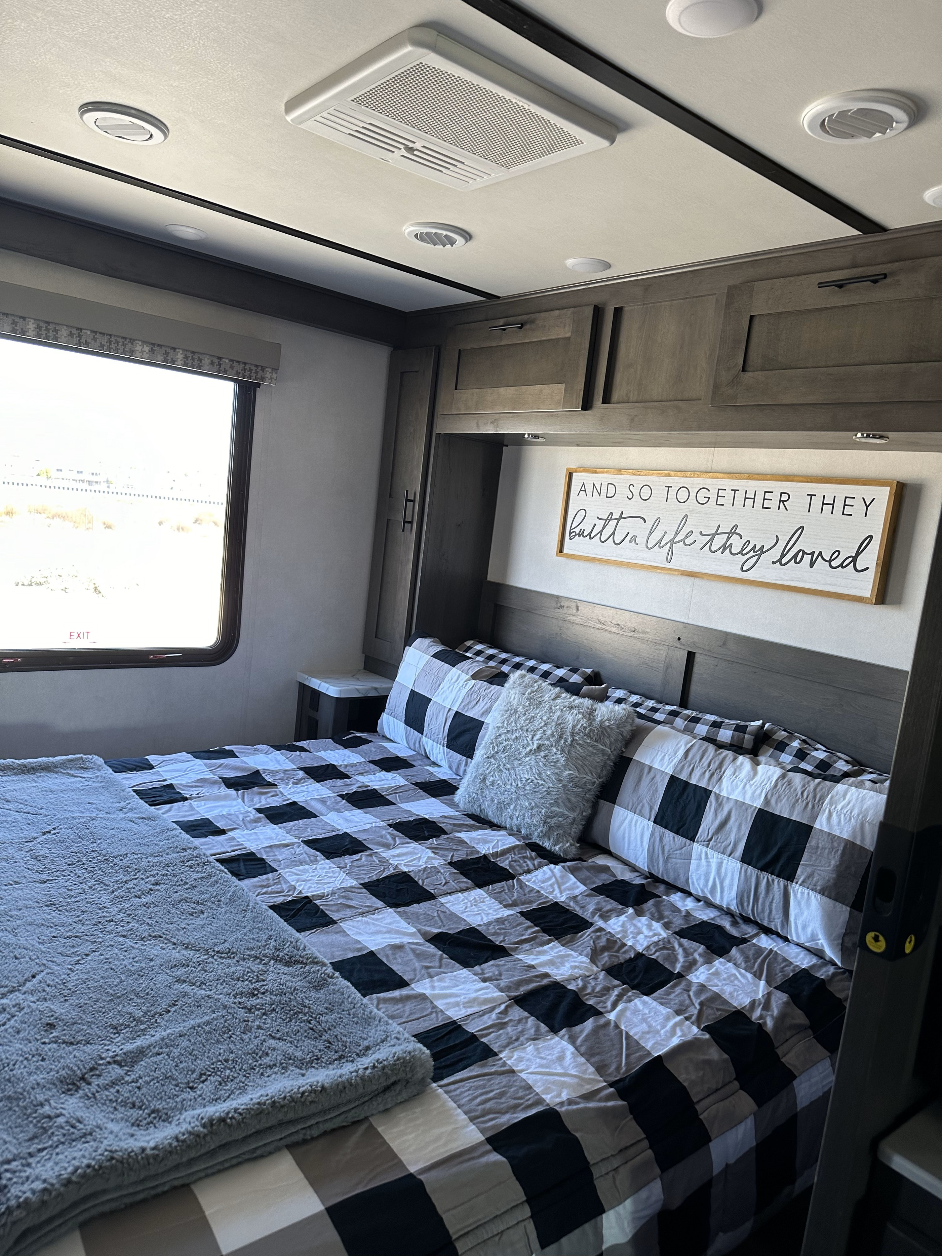 Inside a comfy RV rental bedroom: buffalo plaid bed, dark cabinets, 'loved life' sign, and an EXIT window.. FR3 FR3 Motorhome 2021