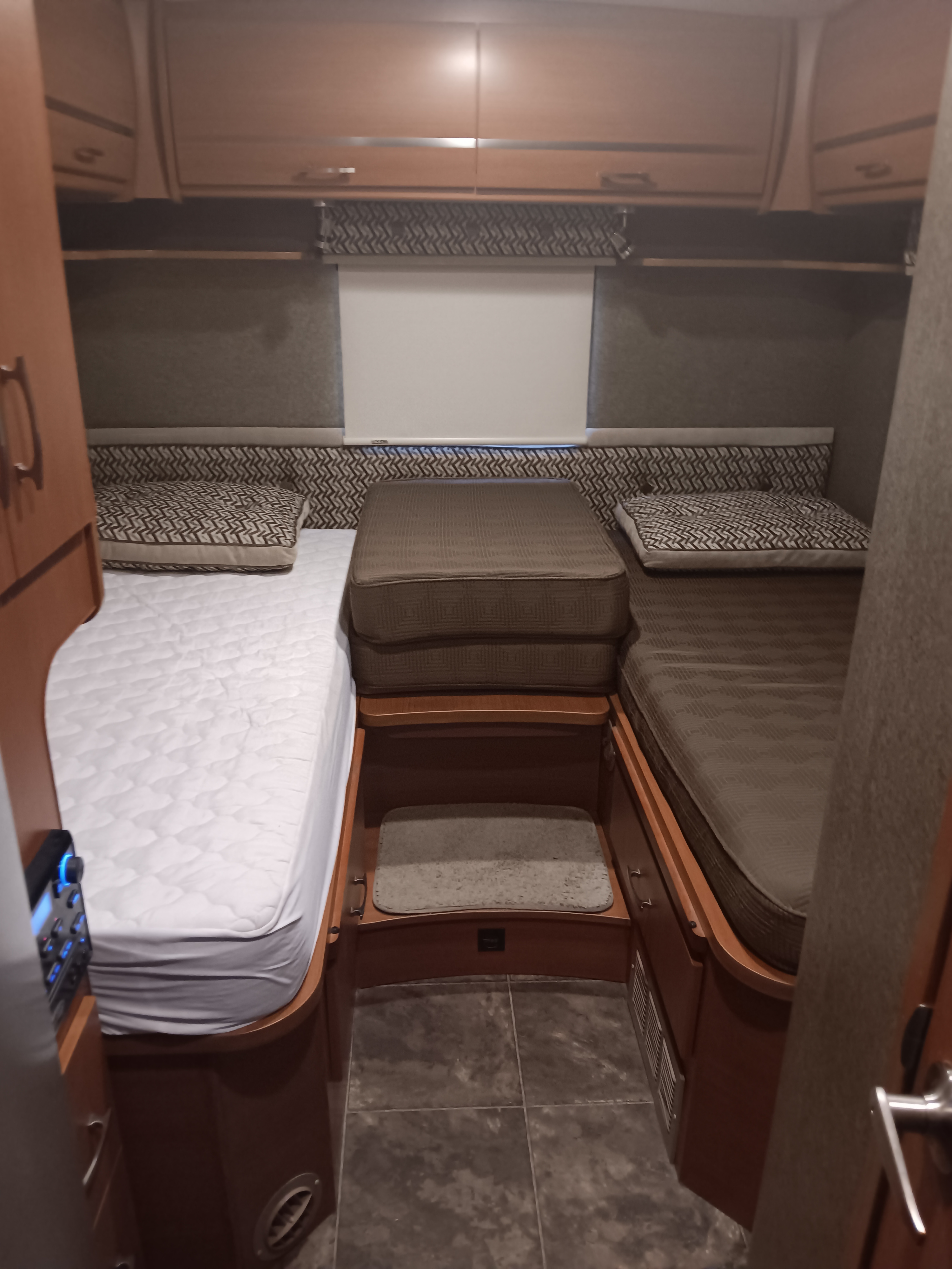 An RV rental's compact bedroom featuring twin beds with patterned cushions, overhead cabinets, and a central aisle.. Itasca Navion 2013