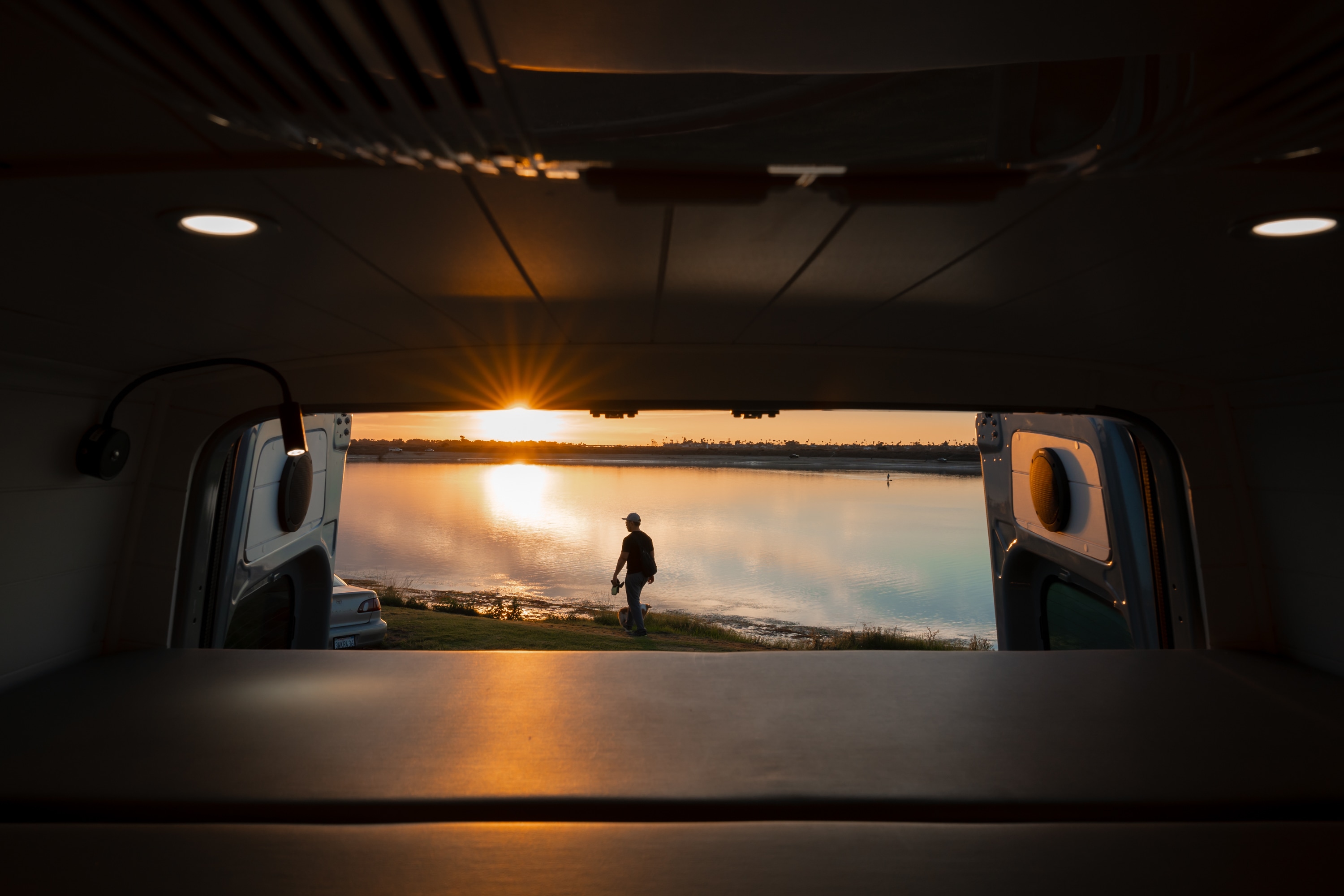 Panoramic sunset from an RV rental. A man & dog stand by shimmering water, seen from inside the cozy van.. Mercedes-Benz Sprinter 2022