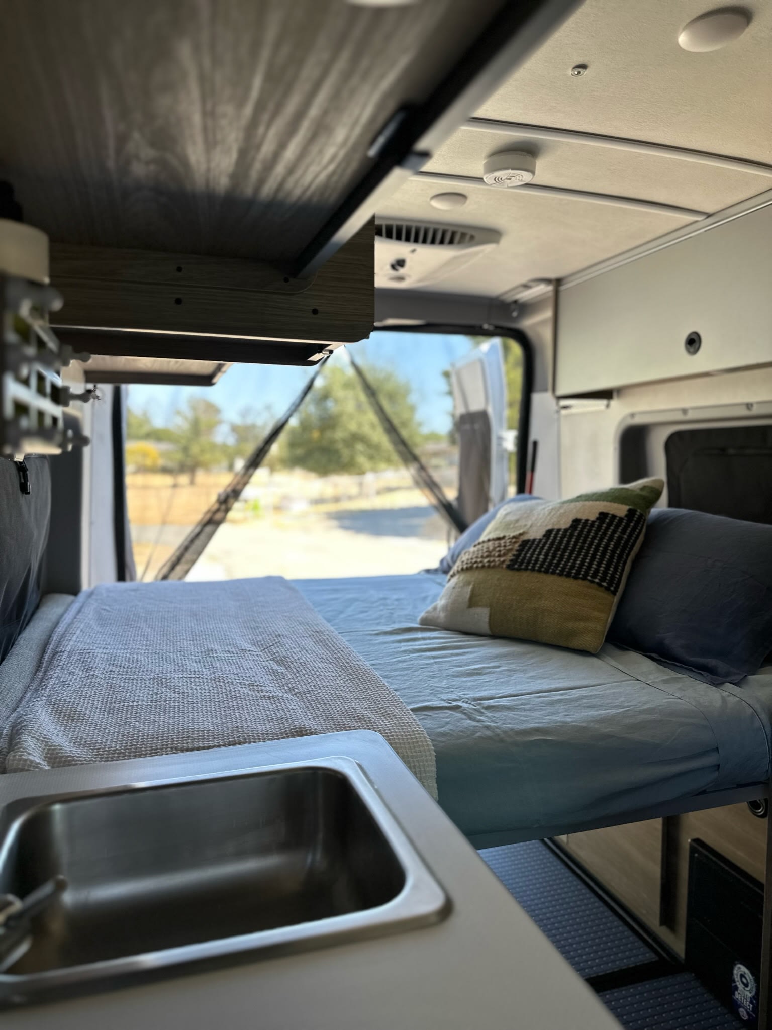 Cozy RV rental interior with a neatly made bed, small sink, and open rear doors revealing an outdoor landscape through a mesh screen.. Winnebago Solis 2023