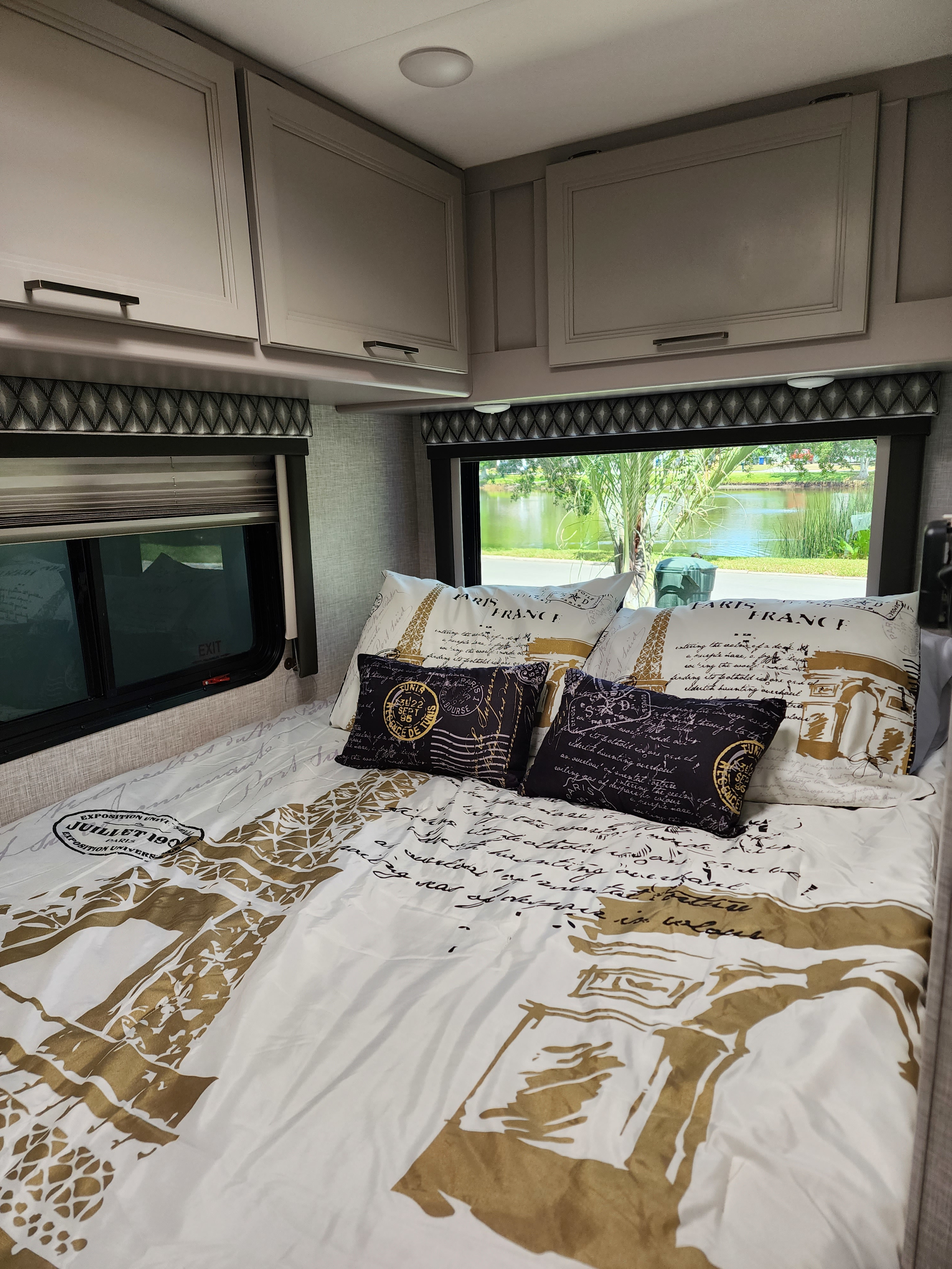 Cozy RV rental bedroom with Paris-themed bedding, overhead cabinets, and a scenic window view of a lake.. Thor Quantum LC 2022