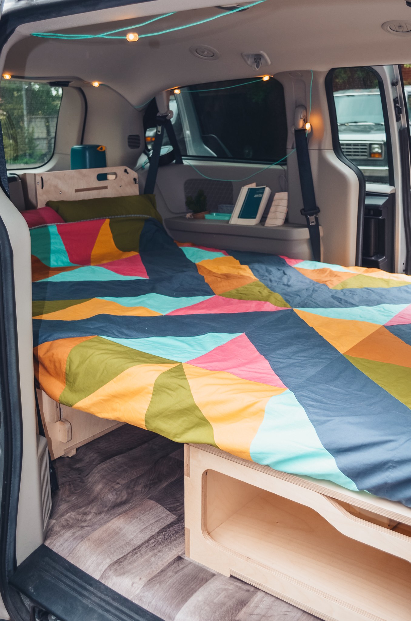 Cozy RV rental interior: a vibrant geometric bed, custom wood storage, and string lights create an inviting campervan space.. Chrysler Grand Caravan 2024
