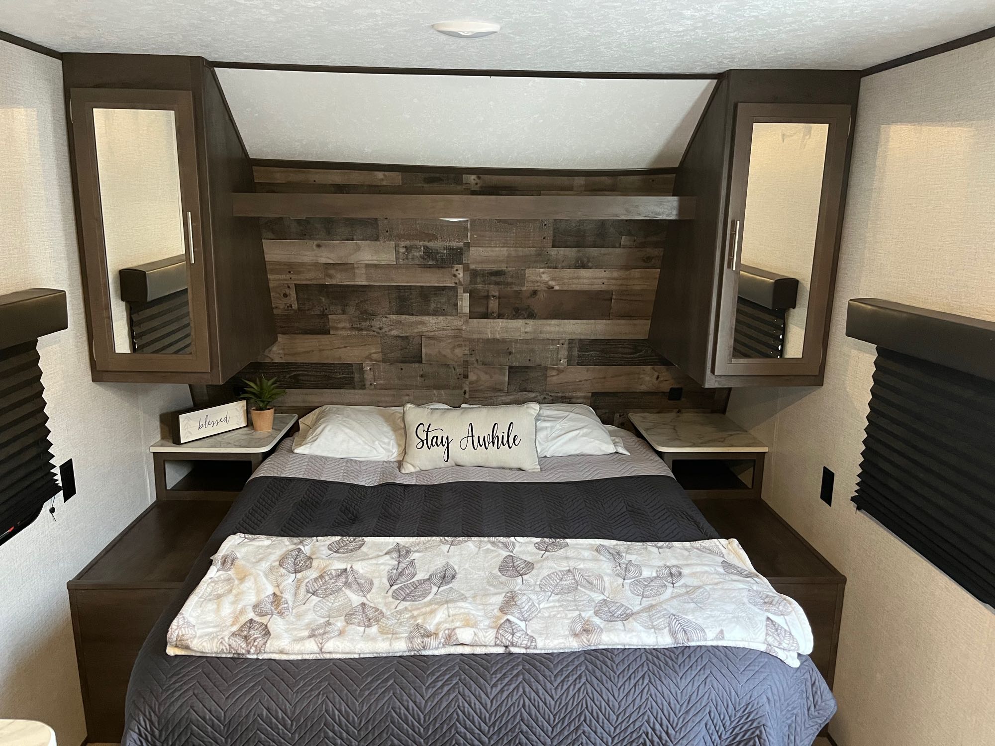 Inside a comfy RV rental bedroom: queen bed, rustic headboard, storage cabinets, and a 'Stay Awhile' pillow.. Dutchmen Coleman Lantern LT 2021
