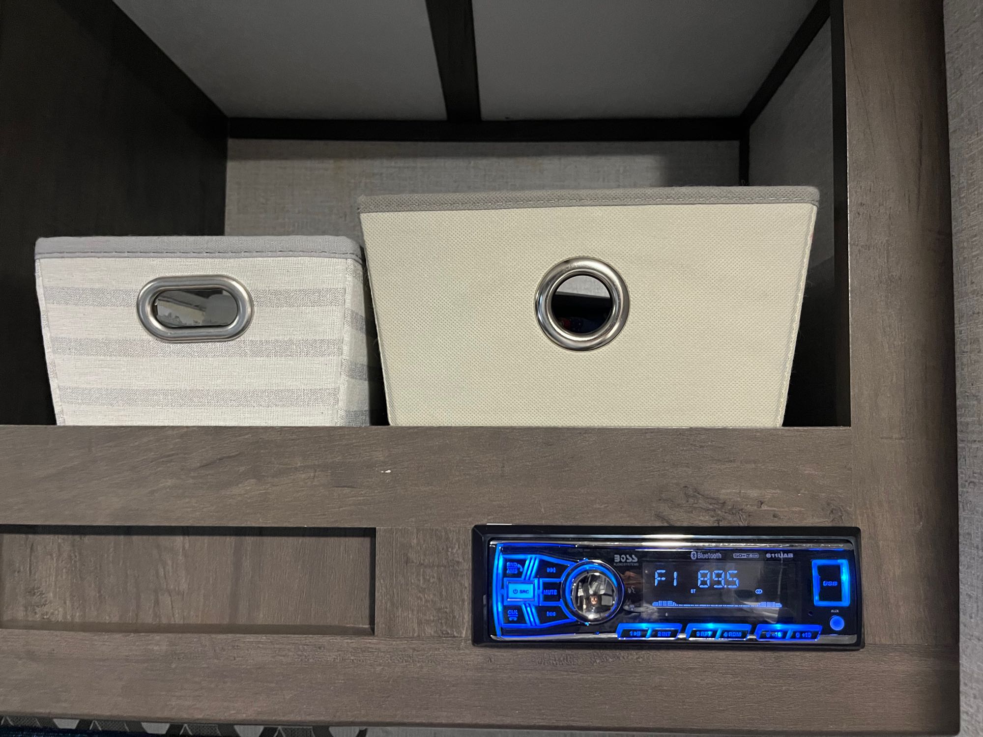 RV rental: Interior view showing two fabric storage bins on shelves above a blue-lit BOSS stereo displaying F1 89.5.. Dutchmen Coleman Lantern LT 2021