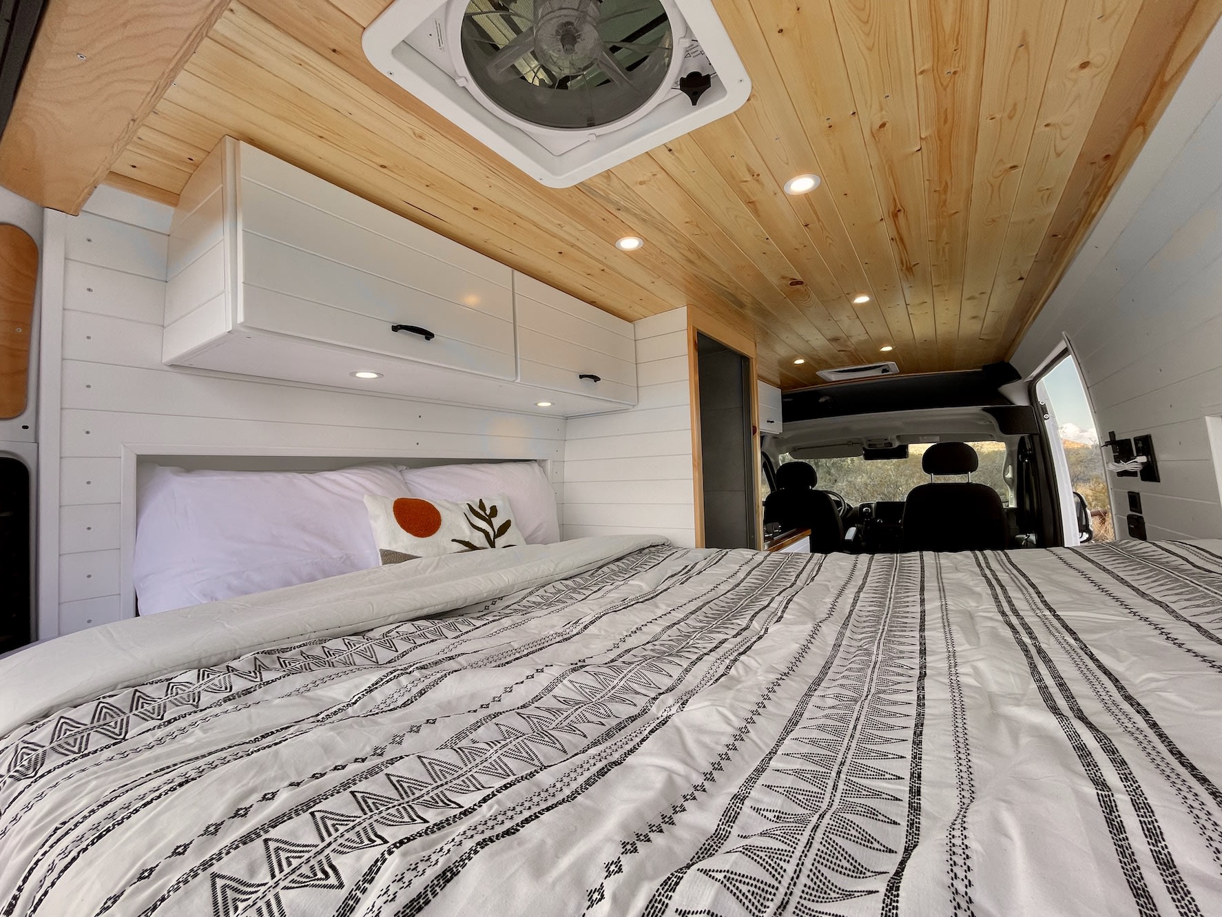 Inside a modern RV rental: view of a comfy bed, white shiplap walls, wood ceiling, & front cab. Ready for travel.. Tommy Campervans Maddox 2023