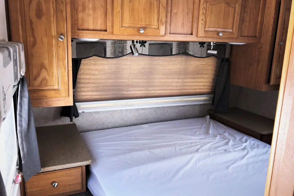 RV rental interior showing a compact bedroom with overhead wood cabinets, bed, and window blinds.. Forest River Rockwood 2007