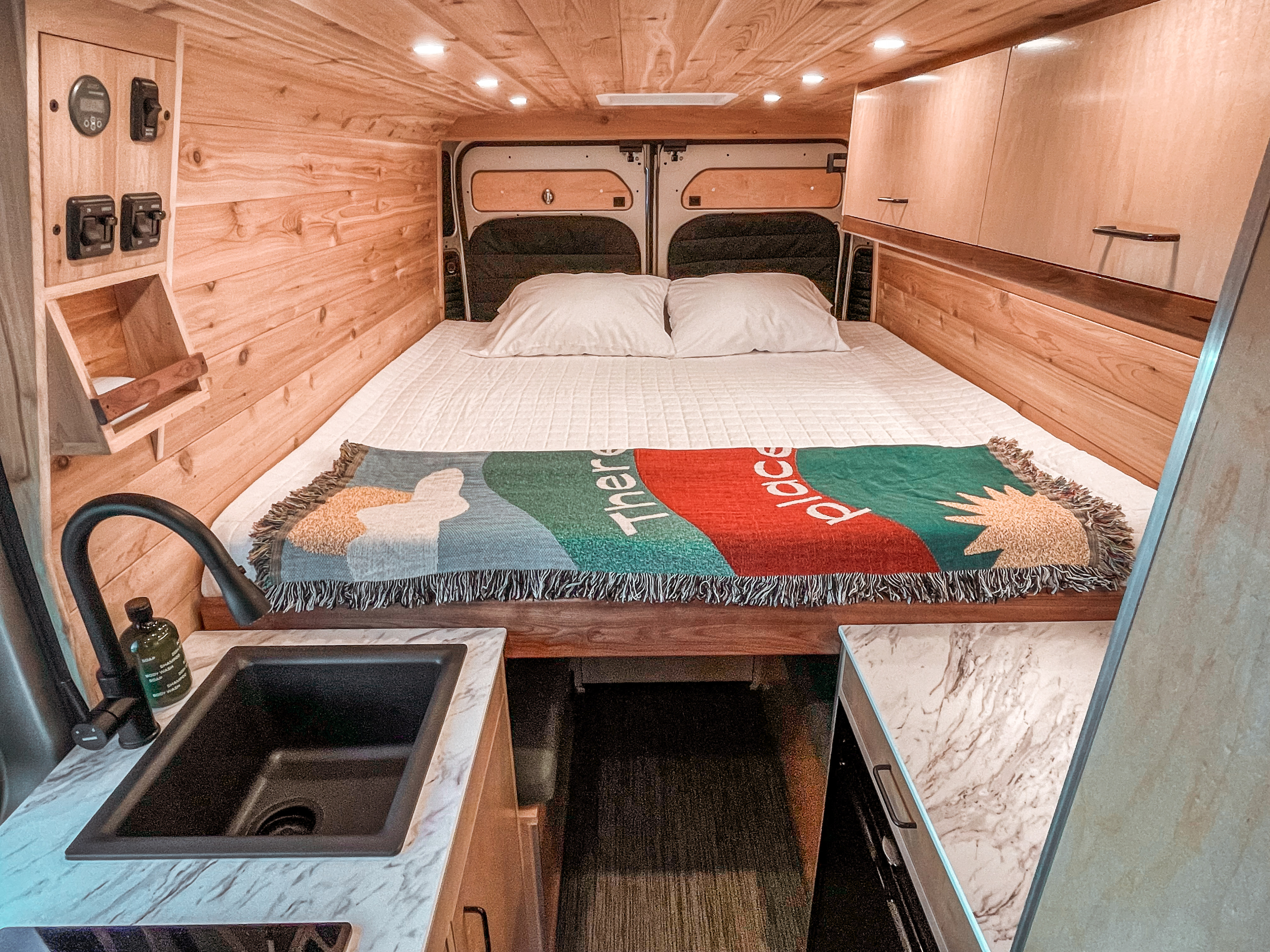 Cozy RV rental interior with a comfortable bed, custom wood paneling, overhead storage, and a functional kitchen sink area.. Ram Promaster 2021