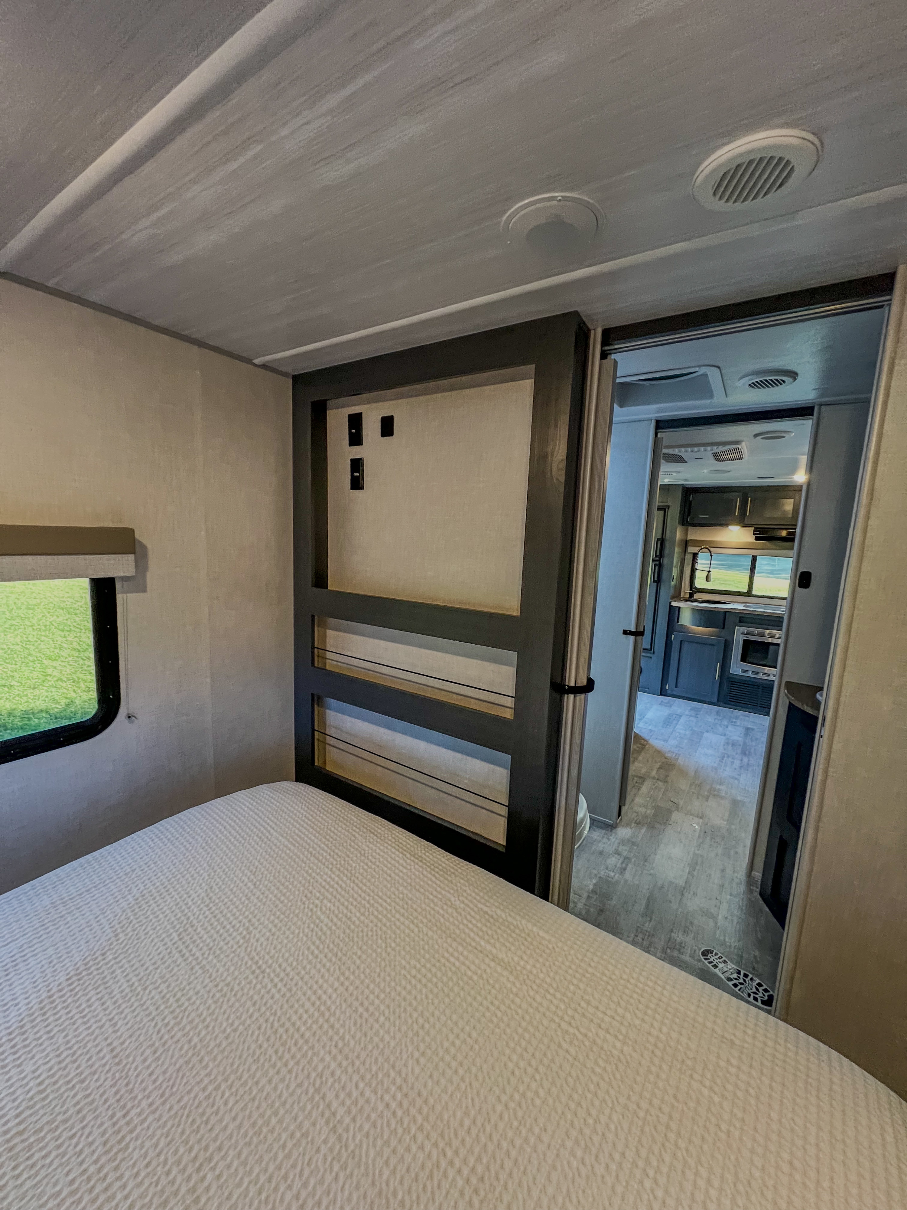 Inside an RV rental, showing a bedroom with a patterned bedspread, and a view through the open door to the kitchen area.. Forest River Rpod 202 2023
