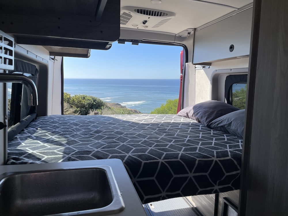 Inside an RV rental: a cozy bed and sink, with stunning ocean views through the open back. Ideal for camper travel.. Winnebago Solis 2023