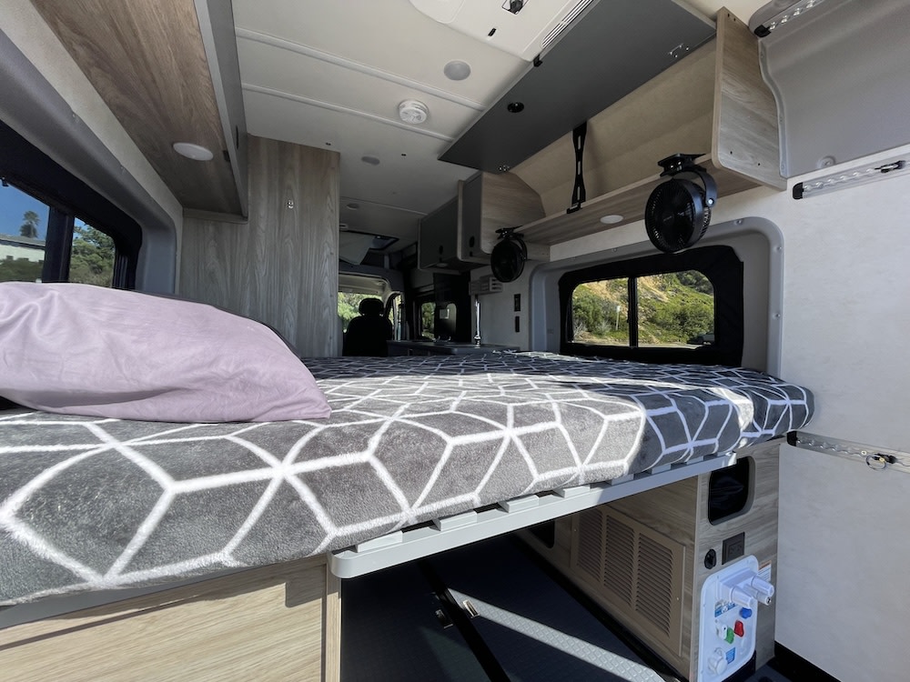 Cozy RV rental interior with a comfortable bed, fans, and scenic window view. Perfect for your adventure.. Winnebago Solis 2023