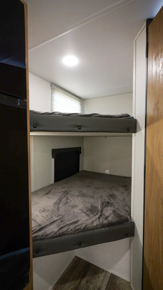 Interior view of an RV rental showing two neatly made bunk beds with grey bedding, a window, and wood-look flooring.. Forest River 19' Bunkhouse with Slide 2024