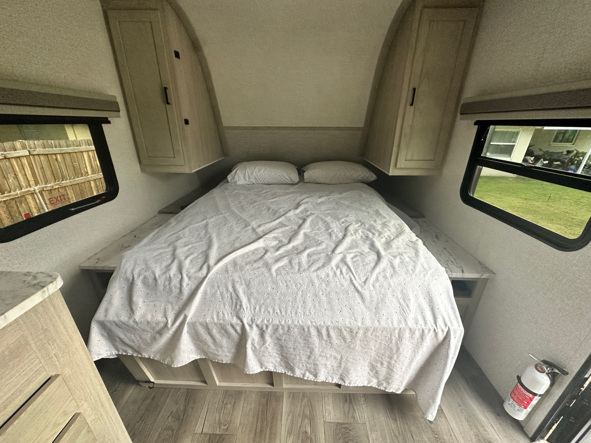This RV rental's compact bedroom features a neatly made bed, overhead storage, and dual windows. Fire extinguisher visible.. East to West Alta 2022
