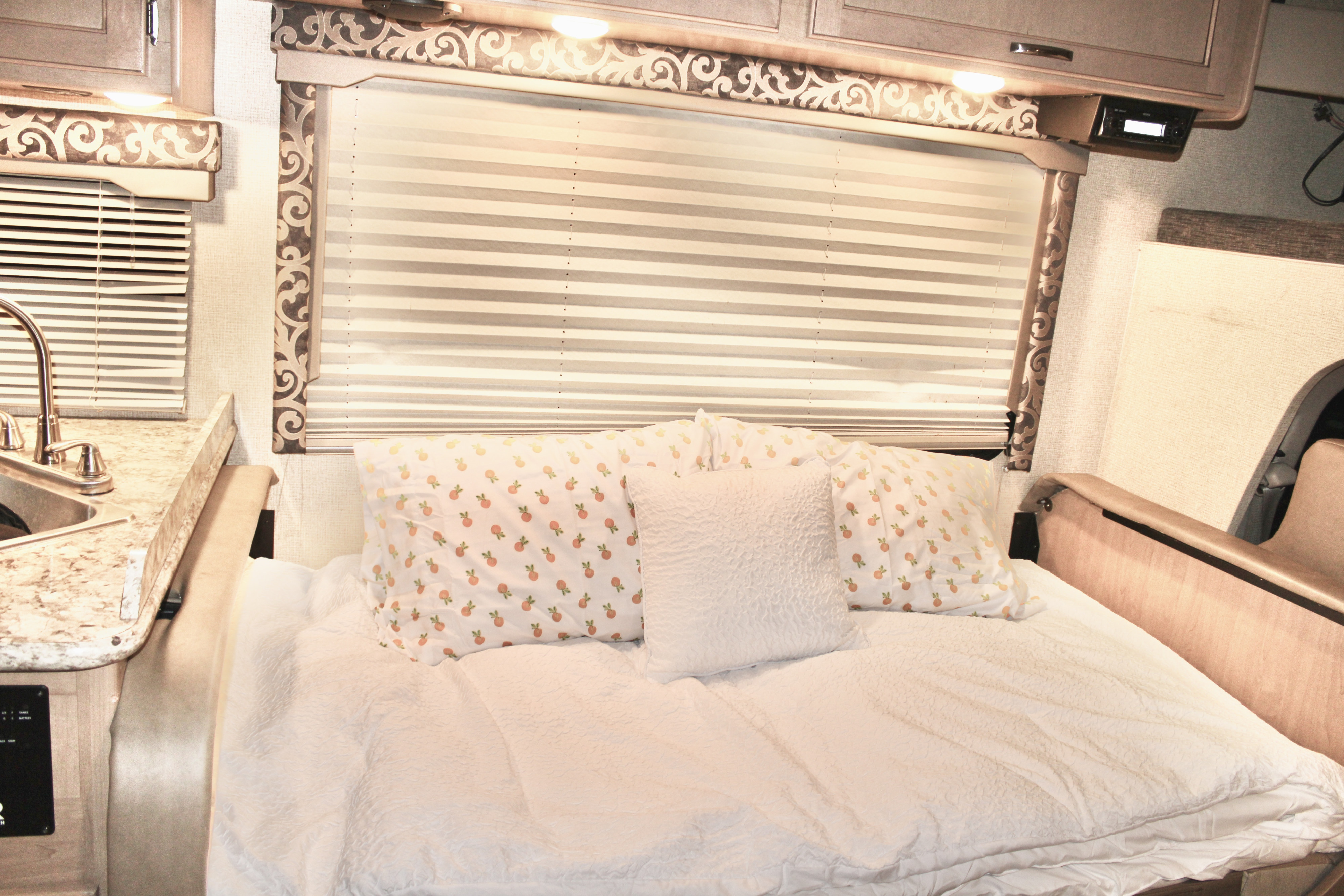 Cozy RV rental interior with a comfortable bed, decorative window blinds, and a compact kitchen area with a sink.. Thor Four Winds 2020