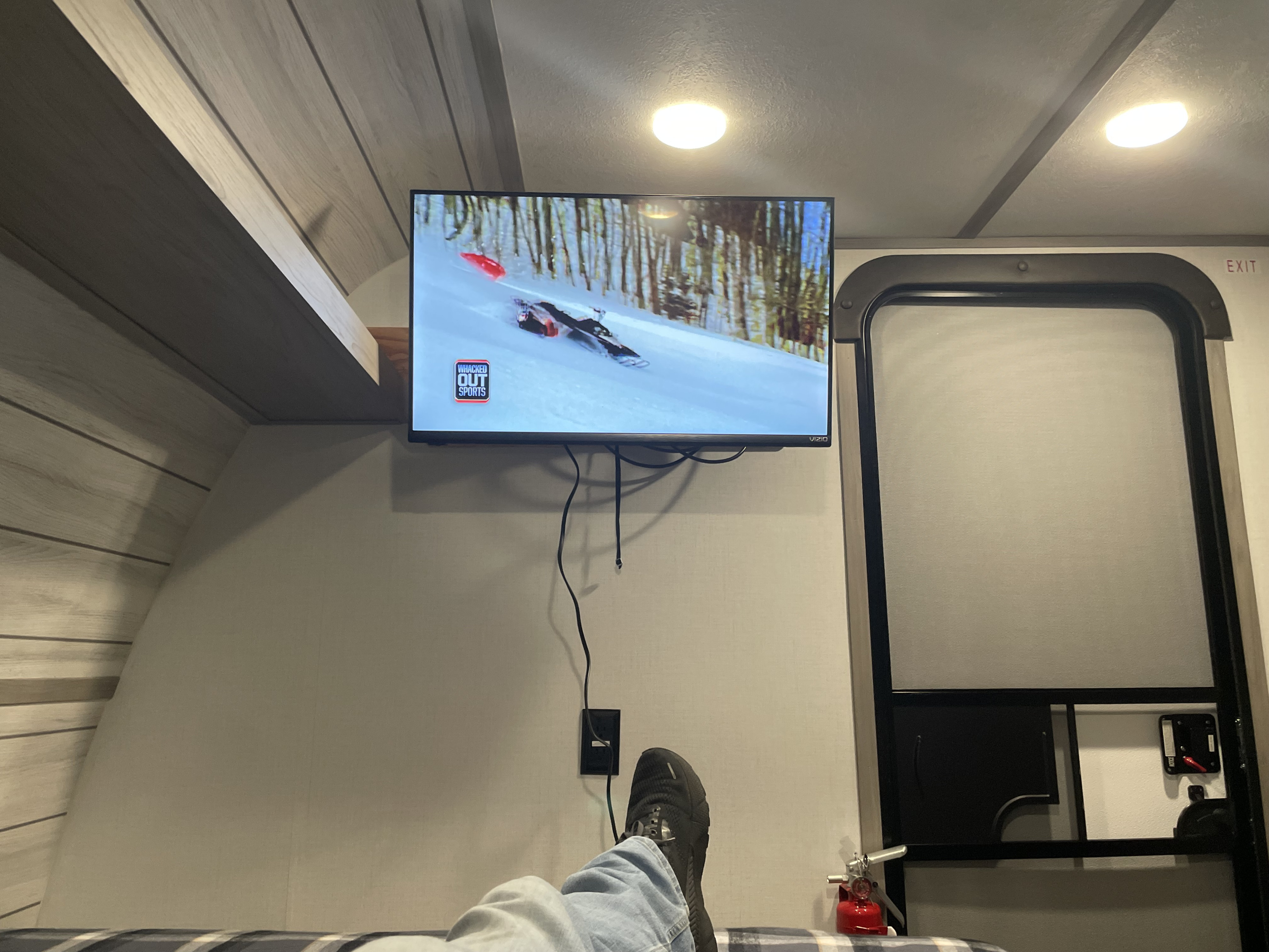 Feet up in an RV rental, watching TV showing snowmobiling. Exit door and fire extinguisher visible.. Coleman 17B 2024