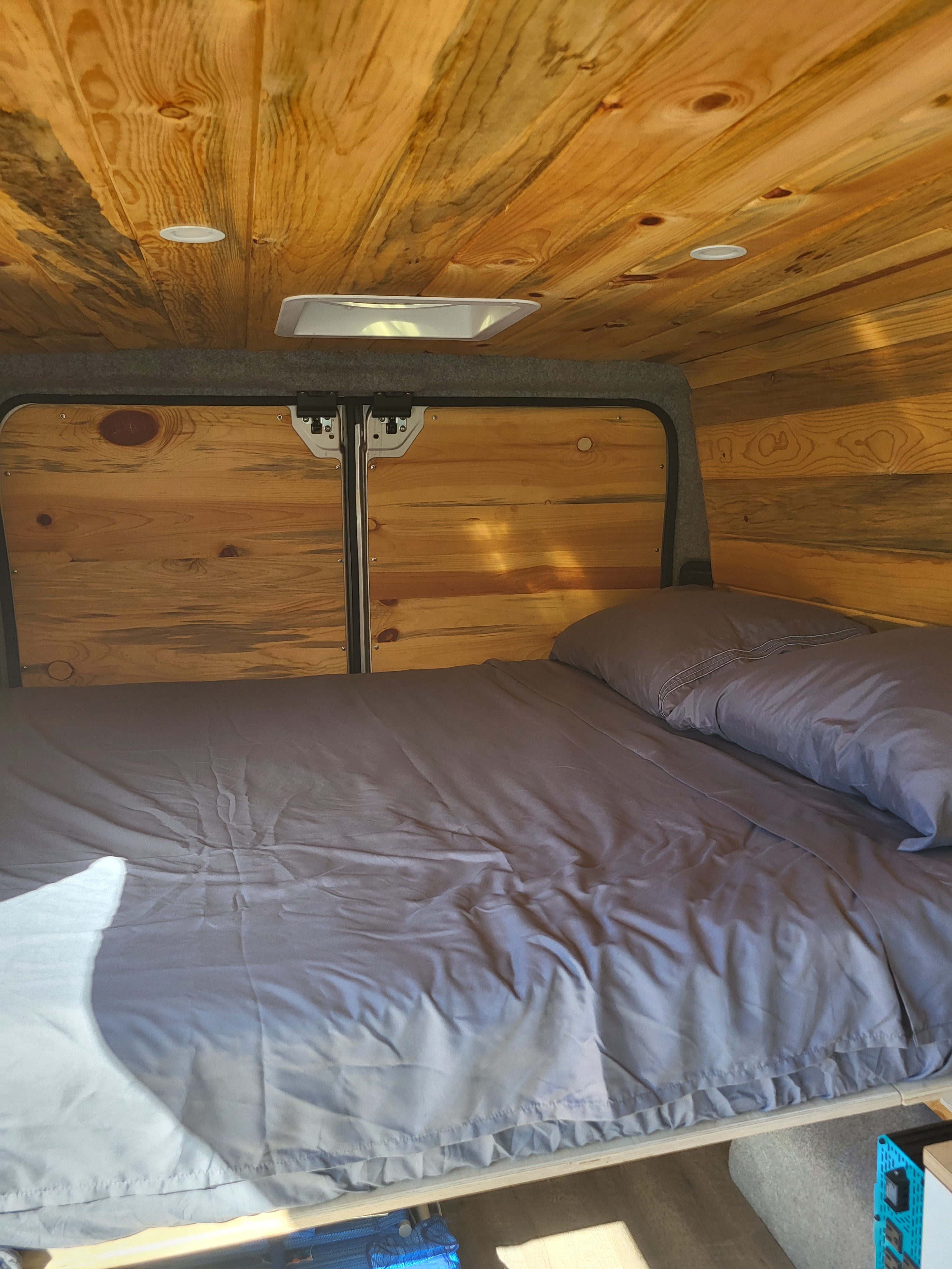Comfortable RV rental bed with a natural wood interior, featuring bright ceiling lights and storage access.. Dodge Ram Promaster 2021