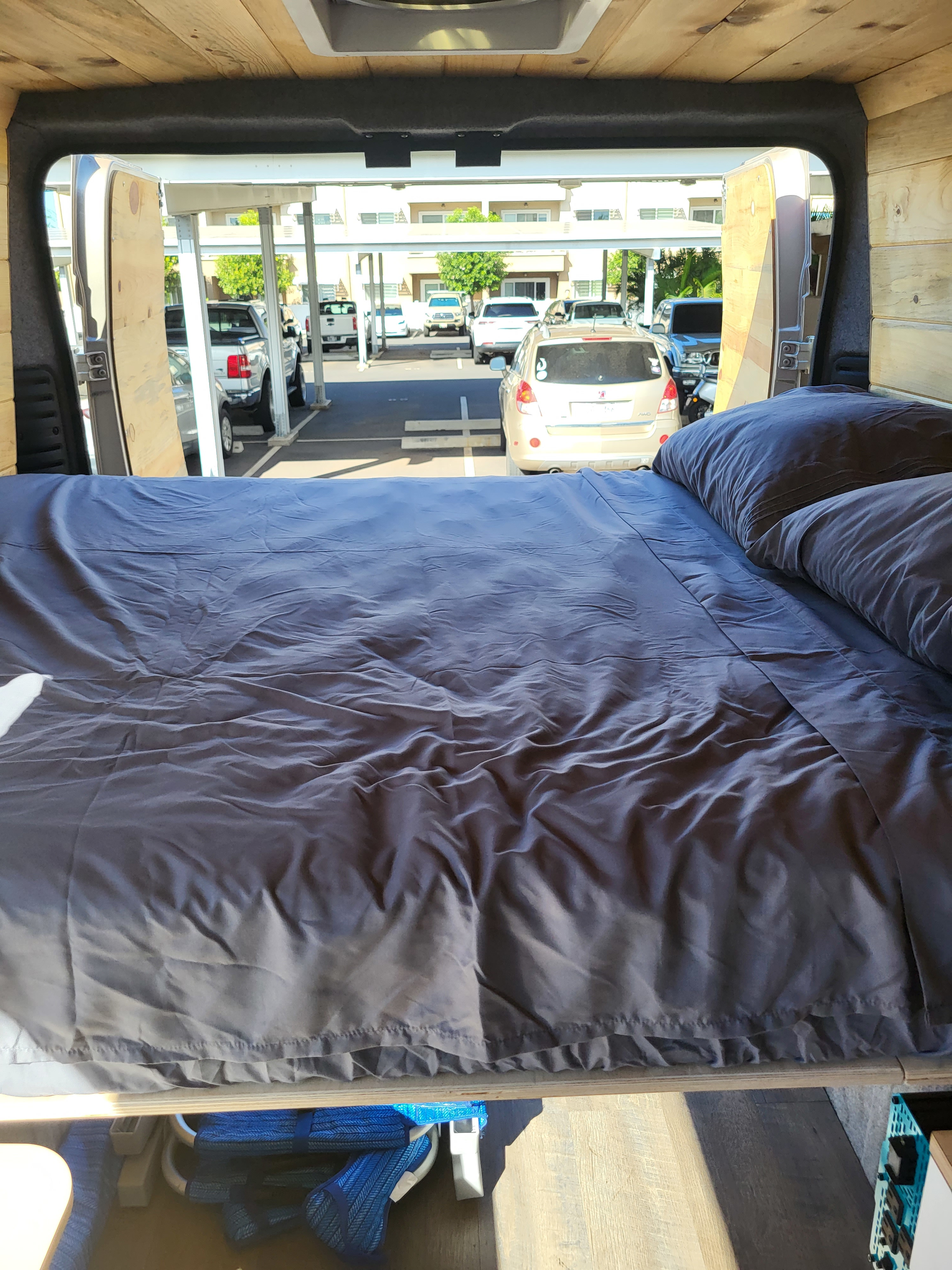 Cozy RV rental interior: a bed with dark bedding, looking out open rear doors to a bright parking lot with cars.. Dodge Ram Promaster 2021