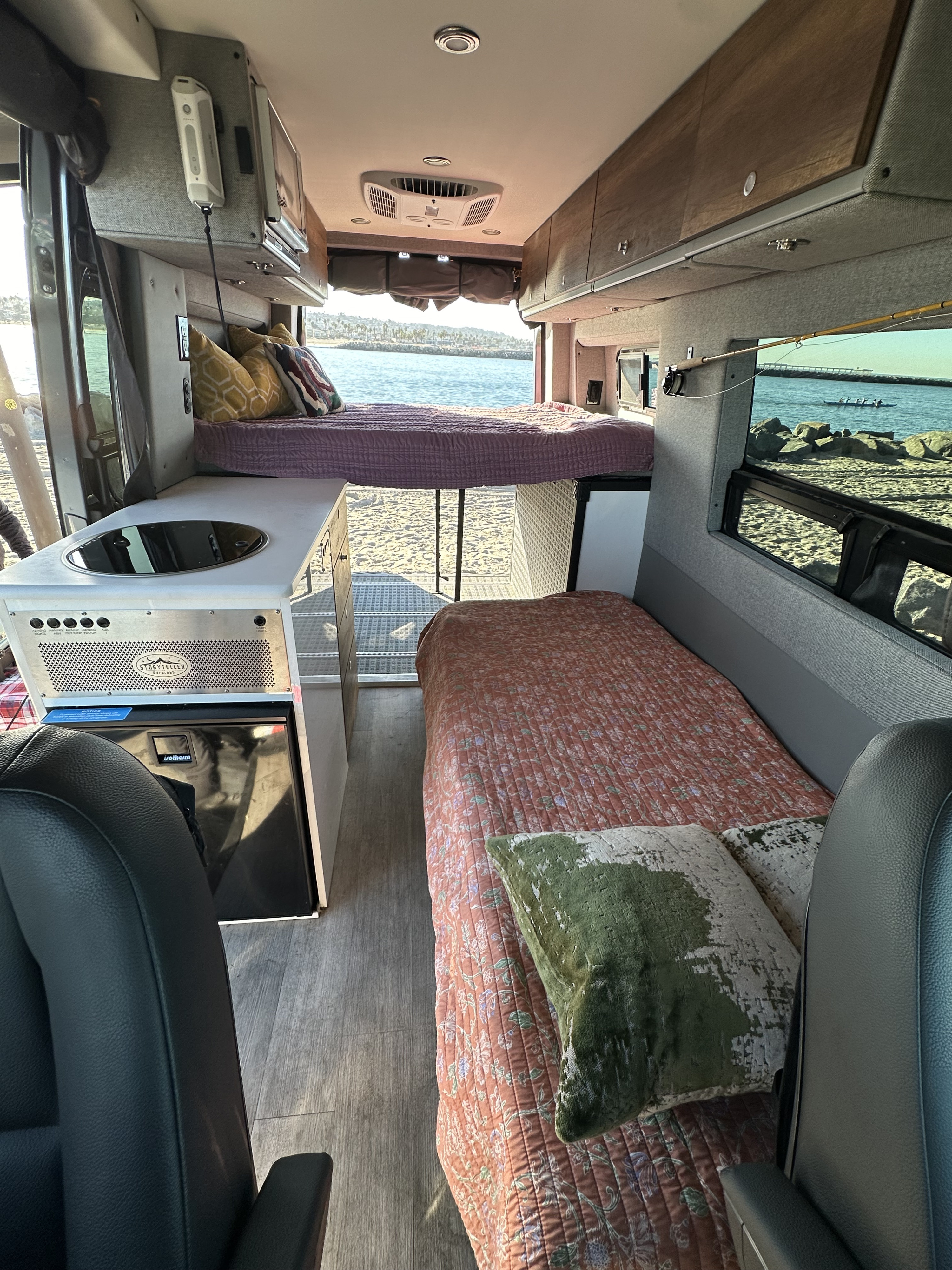 RV rental interior: two beds, kitchenette, and open rear door revealing a beach and ocean view.. Storyteller Overland Storyteller Overland Class B 2023