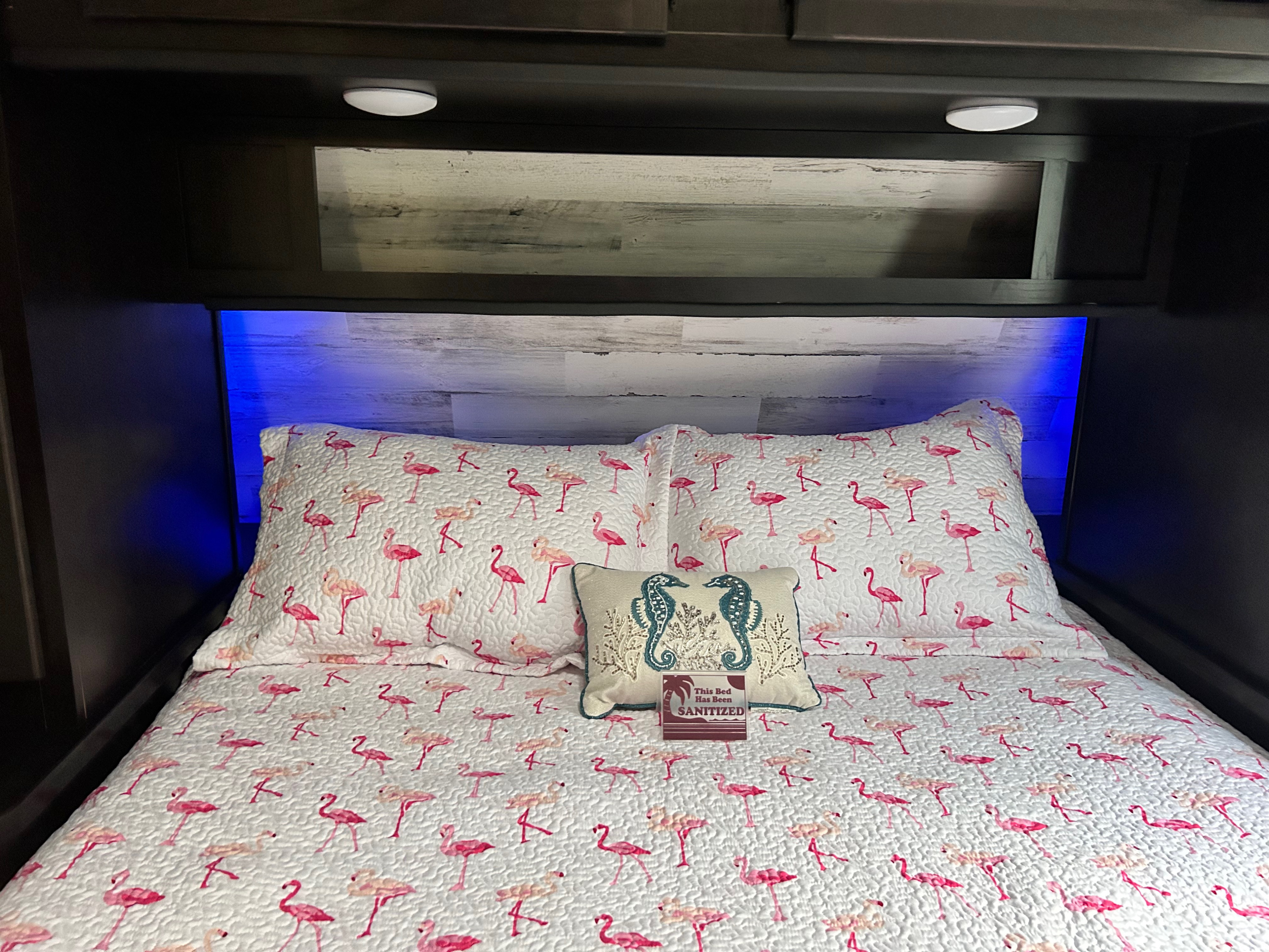 Inviting RV rental bed with whimsical flamingo bedding, seahorse pillow, and blue light. Marked SANITIZED for your travel comfort.. Jayco Jay Feather 2022