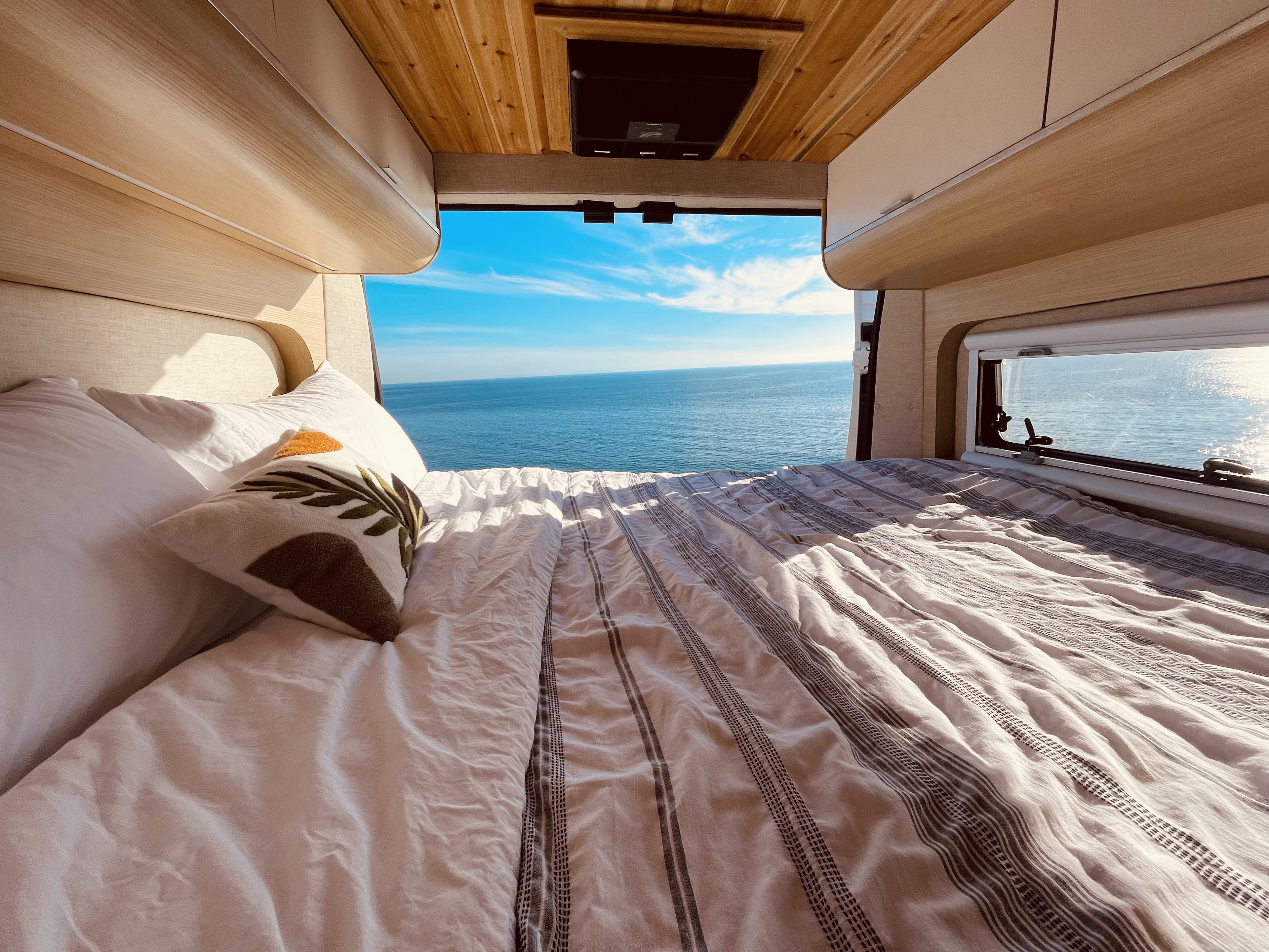 Inside a comfortable rv rental with a made bed looking out to a vast, blue ocean. Ideal for serene camper van travel.. Noovo Lite 2023