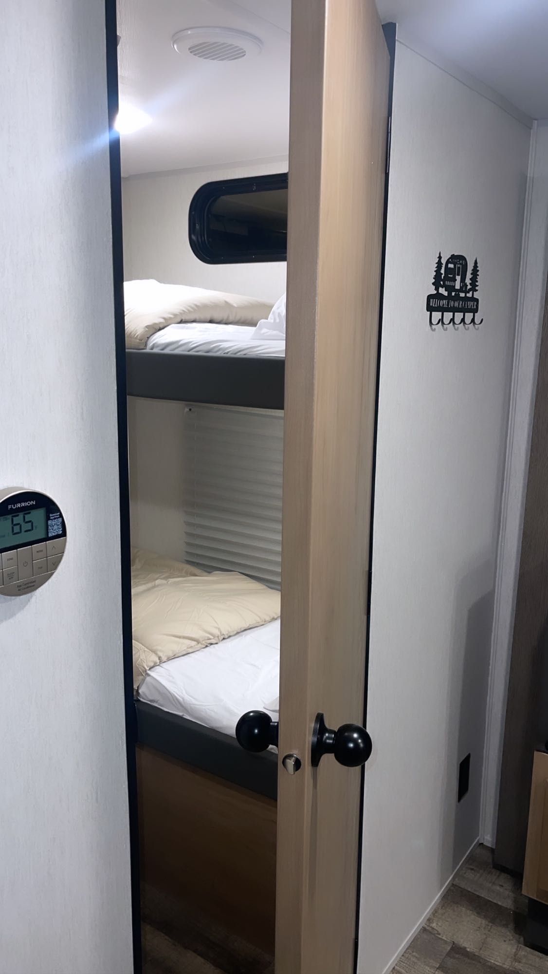 Inside an RV rental, cozy bunk beds are visible past an open door. A thermostat reads 65, and a "Welcome to our camper" sign hangs.. Forest River Cherokee Grey Wolf 29TE 2024