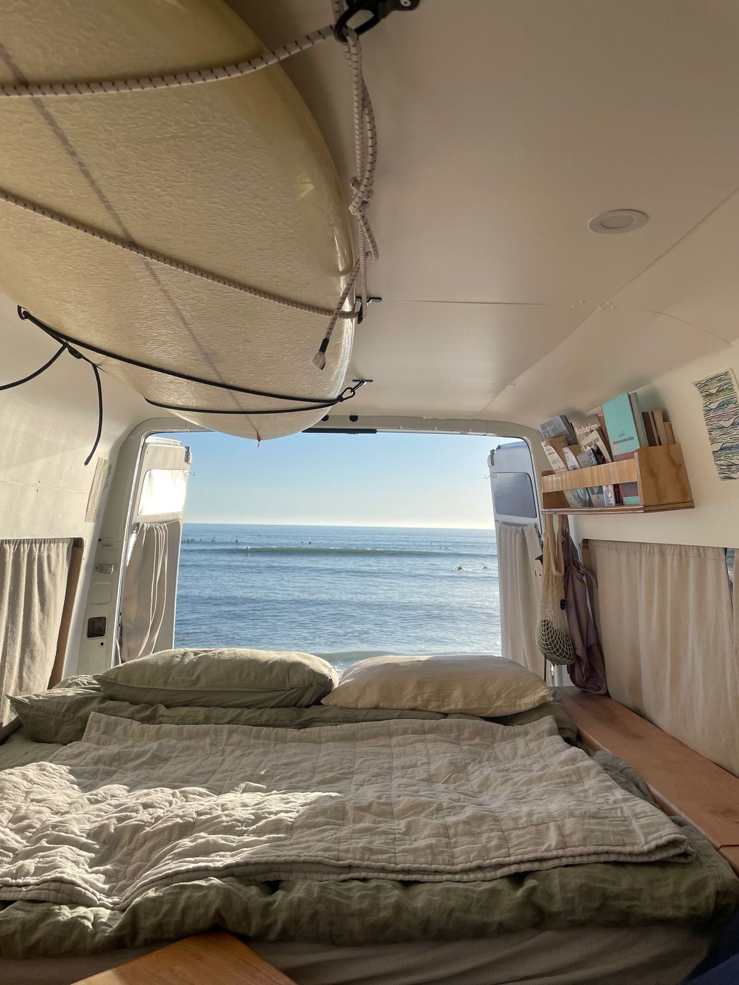 Inside an RV rental: a cozy bed faces an ocean view with surfers. A surfboard is stored above.. Dodge Sprinter Wagon 2500 2003