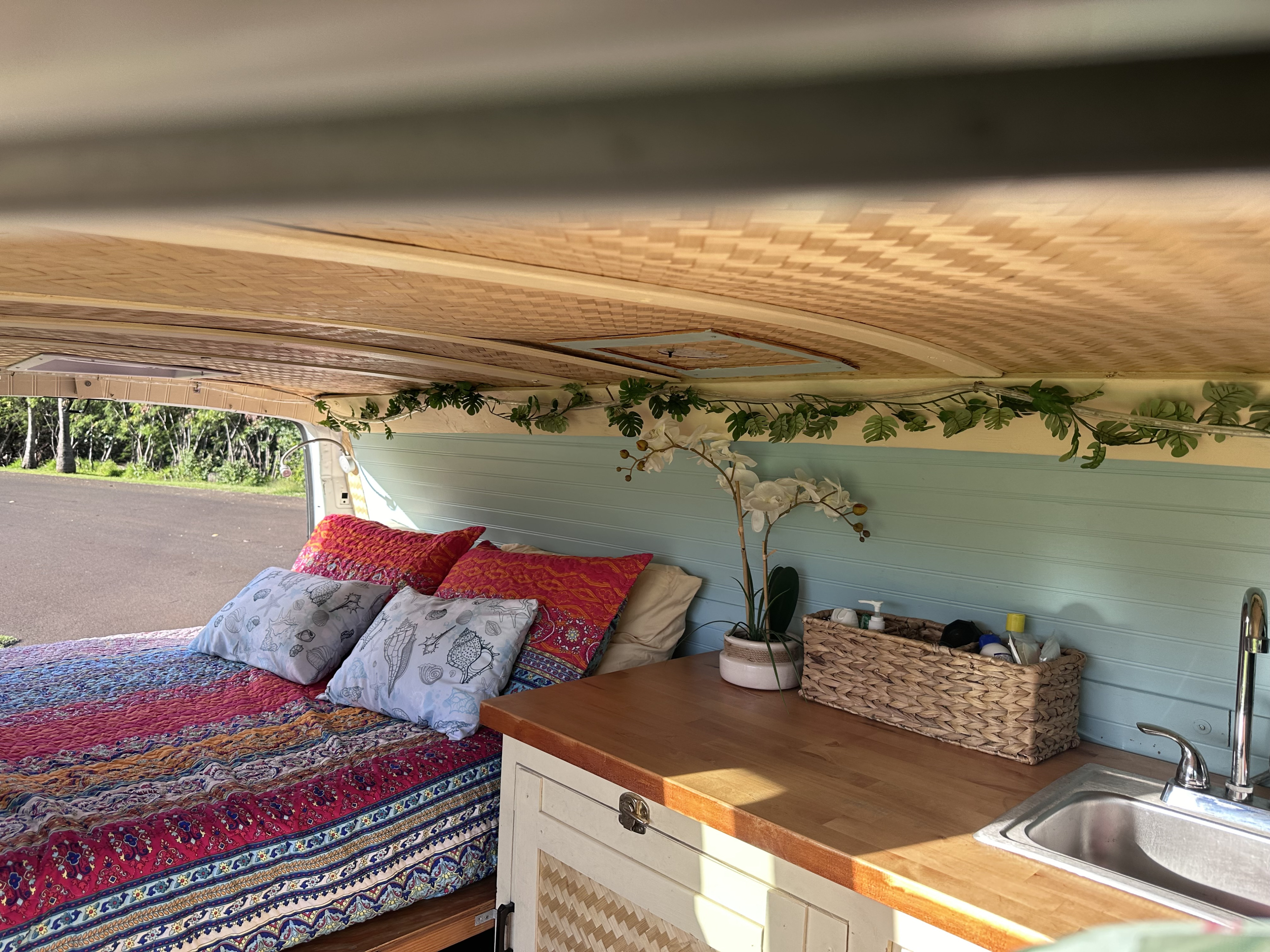 Cozy RV rental interior: colorful bed, mini kitchen with sink, light blue shiplap walls, and woven ceiling. View outside open door.. Gmc Savanna 2006